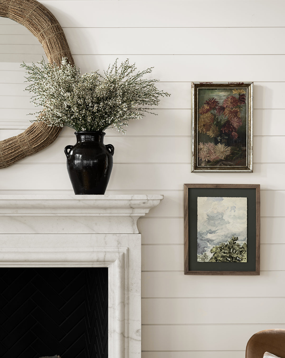 A white shiplap wall features a black vase with white flowers on a mantel, a round woven mirror, and two framed paintings—a floral print and Head in the Clouds, an art landscape of a cloudy day.