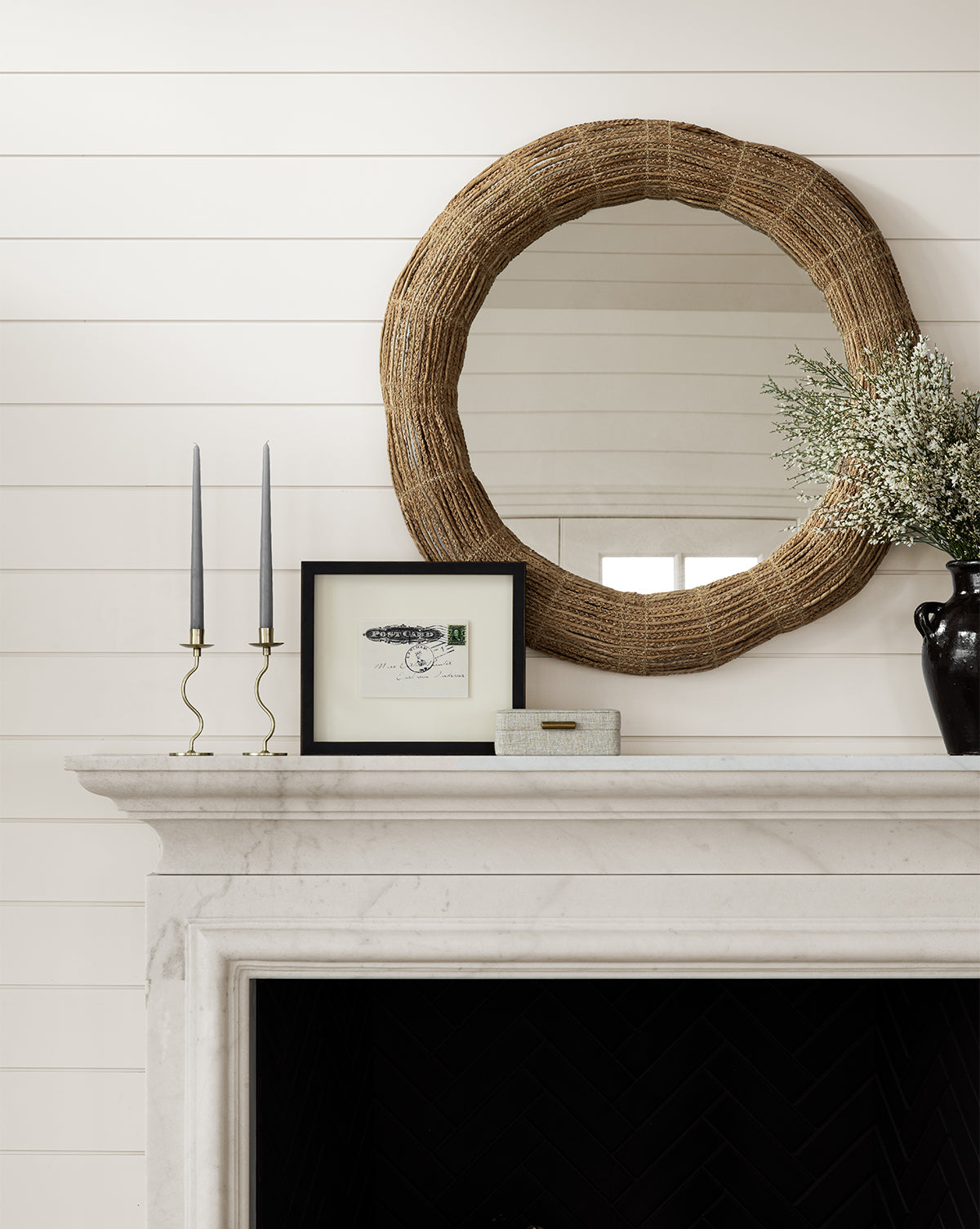 A white mantel features a black vase with flowers, two tall gray candles in gold holders, a small framed picture, a decorative box, and the Gareth Woven Mirror above on a shiplap wall by McGee & Co.