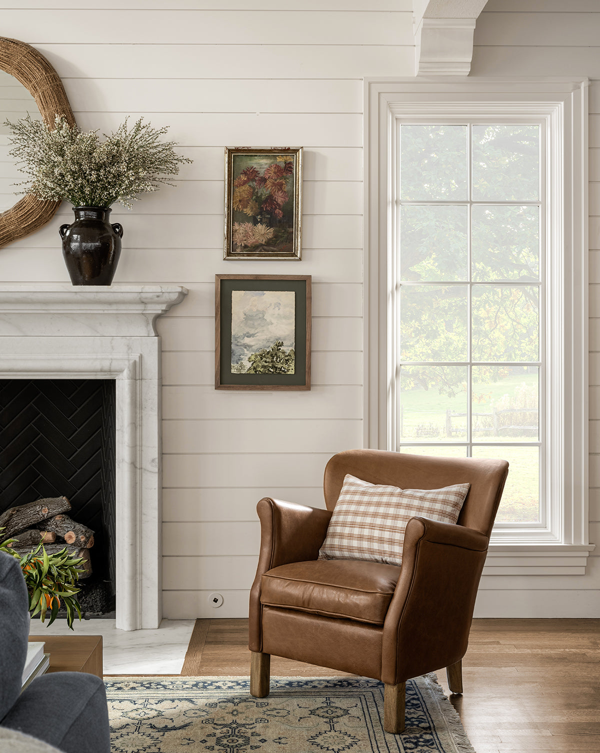 A cozy living room corner features the Jarah Lounge Chair in top-grain leather, a plaid pillow, marble fireplace with a vase of flowers, two paintings on a shiplap wall, and natural light from a large window, by McGee & Co.
