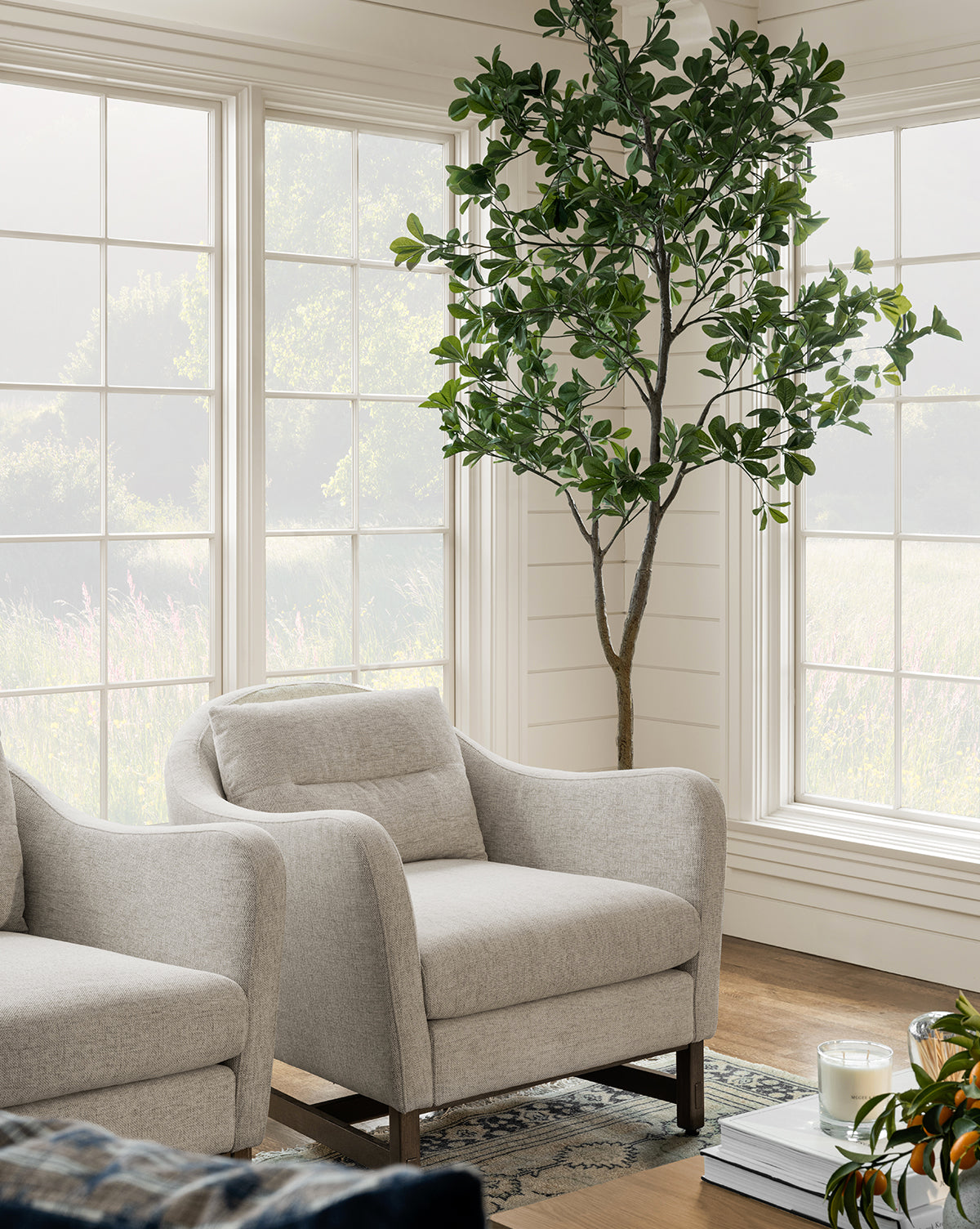 A bright living room with large windows features a Shady Lady Faux Tree in a ceramic pot, two light gray armchairs on a patterned rug, and a small table topped with a candle and plant.