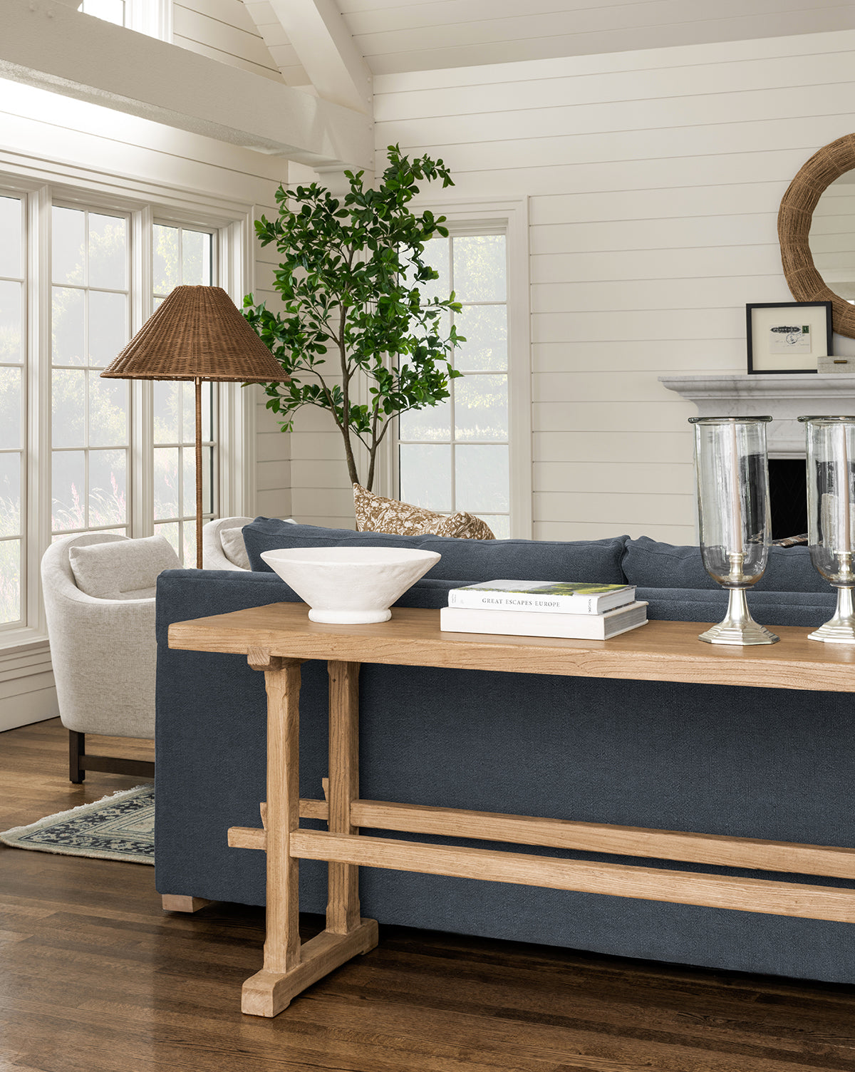A bright living room with large windows, a dark blue sofa, a wooden console table, the Matias Bowl, books, two glass candle holders, a potted plant, a round mirror, and a white armchair by the fireplace.