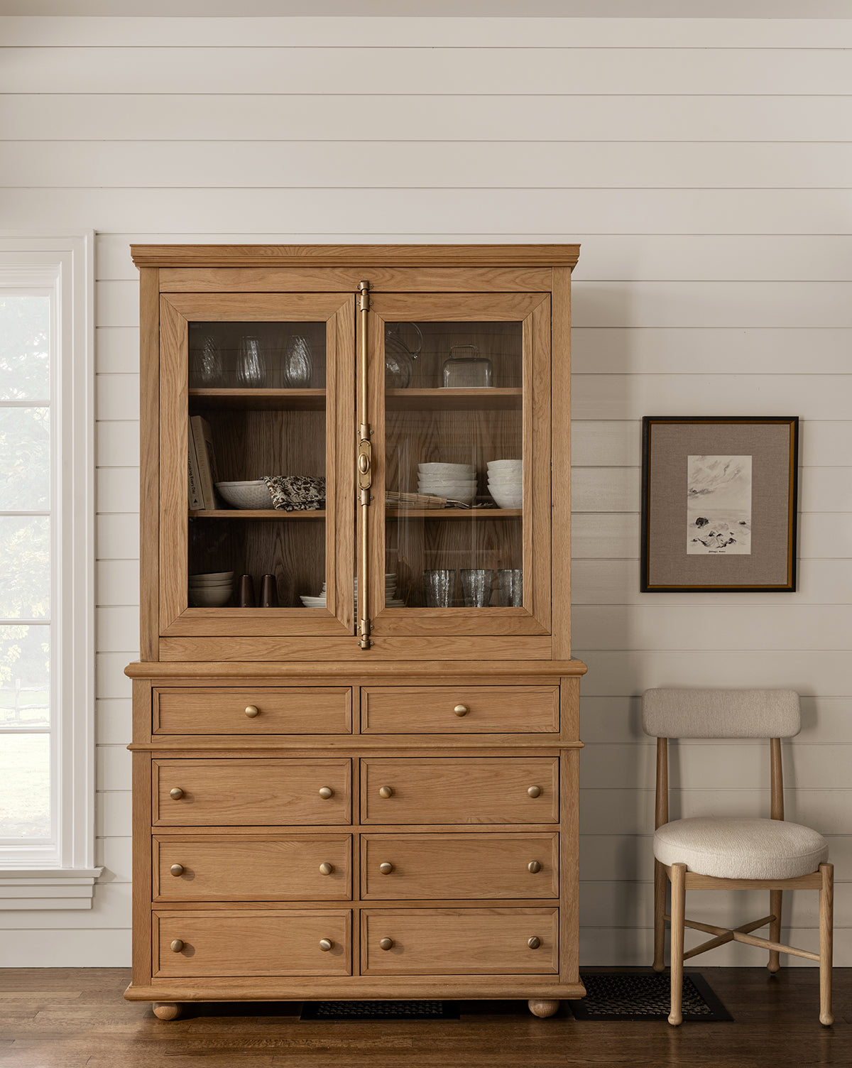 A cushioned chair sits beside a wooden hutch with glass doors and drawers, while “September in Kalamata,” a giclee print with hand-wrapped linen mat in a distressed black frame, hangs on the white shiplap wall by the window.