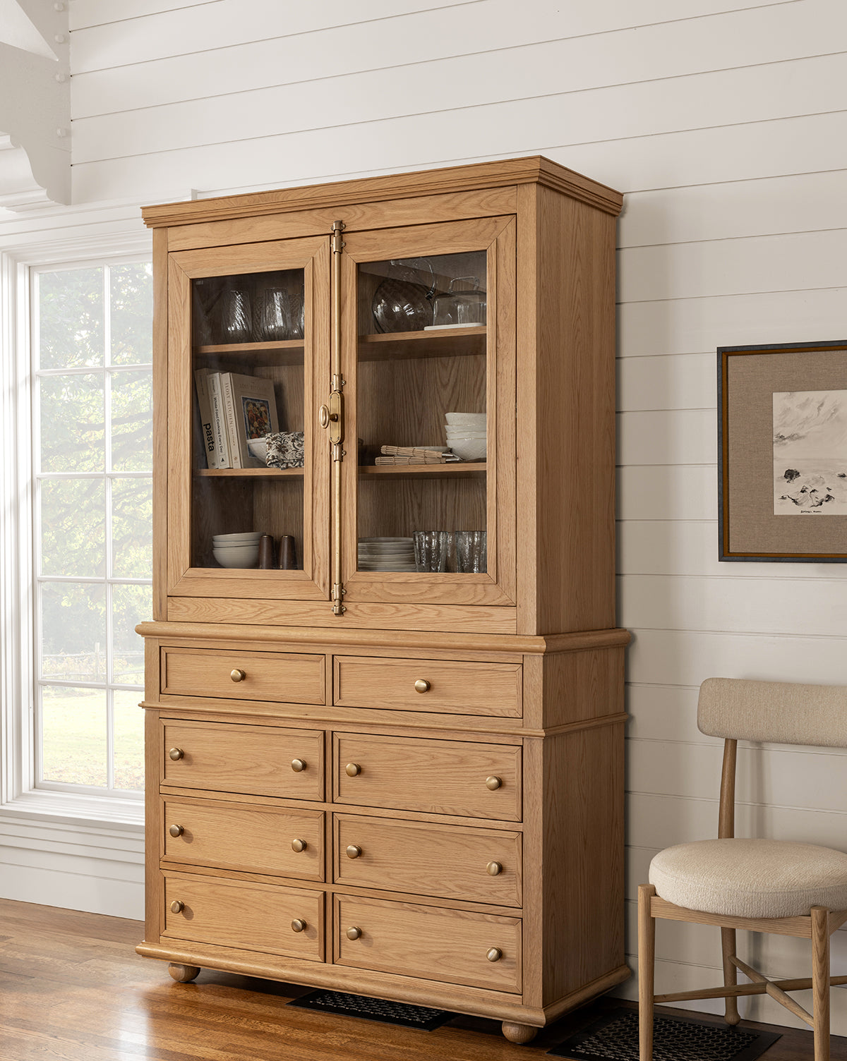 The Heath Oak Cabinet features glass doors above to showcase glassware and dishes, with eight round-knob drawers below. Set near a window against a white shiplap wall and beige chair, it highlights timeless traditional design by McGee & Co.