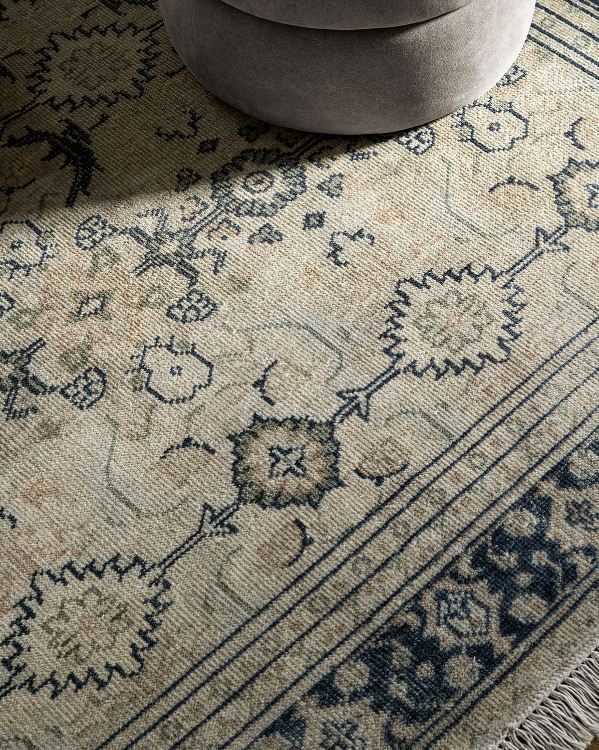 A close-up of the Vercelli Hand-Knotted Wool Rug, showing its intricate blue and beige patterns with tasseled edges, alongside part of a round light gray upholstered ottoman in the upper corner, by McGee & Co.