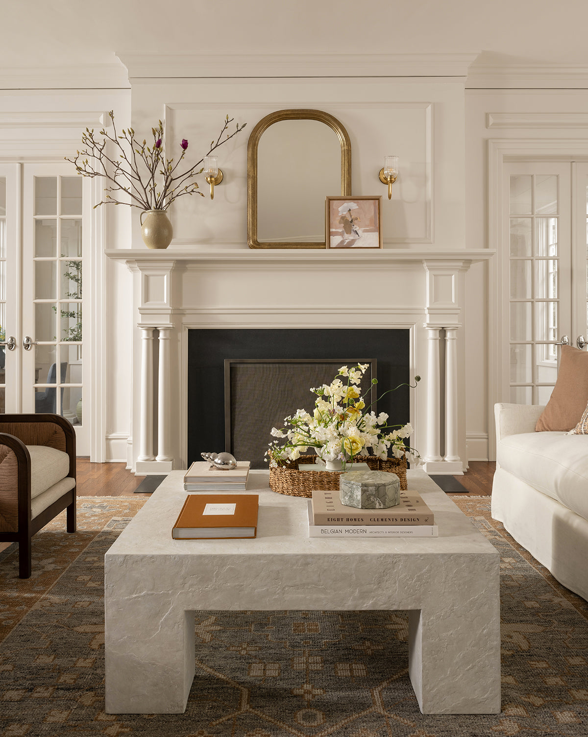 A bright, elegant living room with a stone coffee table, floral arrangements, books, a white sofa and armchair, and a white fireplace featuring the Giada Wall Mirror in its moulded frame with decorative objects above the mantel, by McGee & Co.