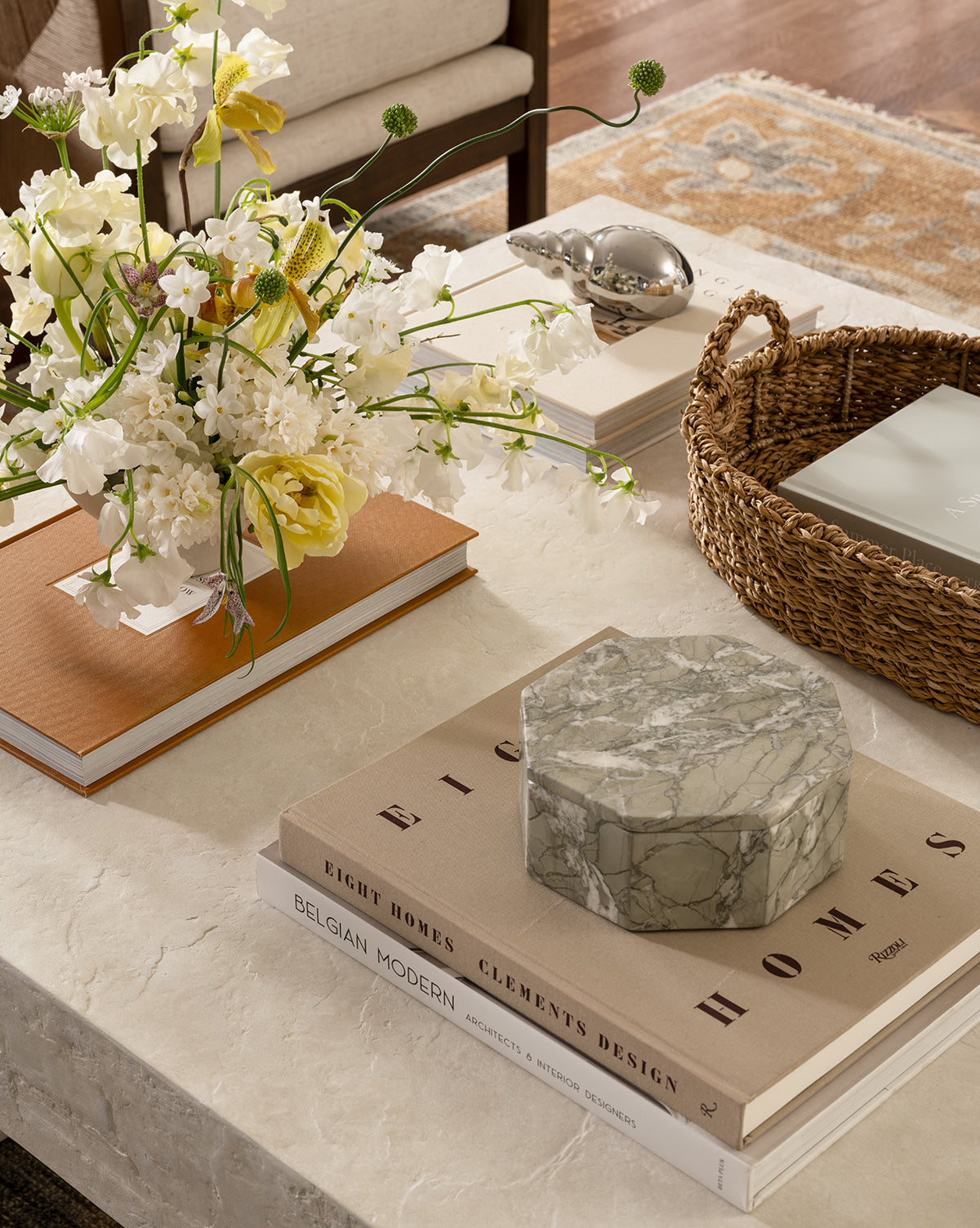 A marble coffee table displays design books, a marble box, a Seagrass Oval Tray with natural accents, white and yellow flowers, and a silver bird sculpture, creating a cozy living room scene.
