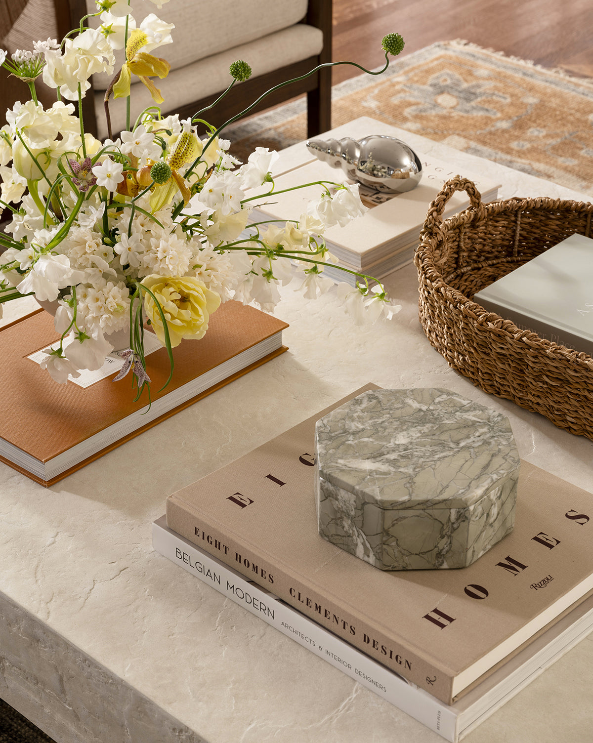 A marble coffee table holds a Paolo Marble Box, stacked books, a woven basket, and a glass bird sculpture. A vase of yellow and white flowers sits on a book. An ornate rug is visible in the background.