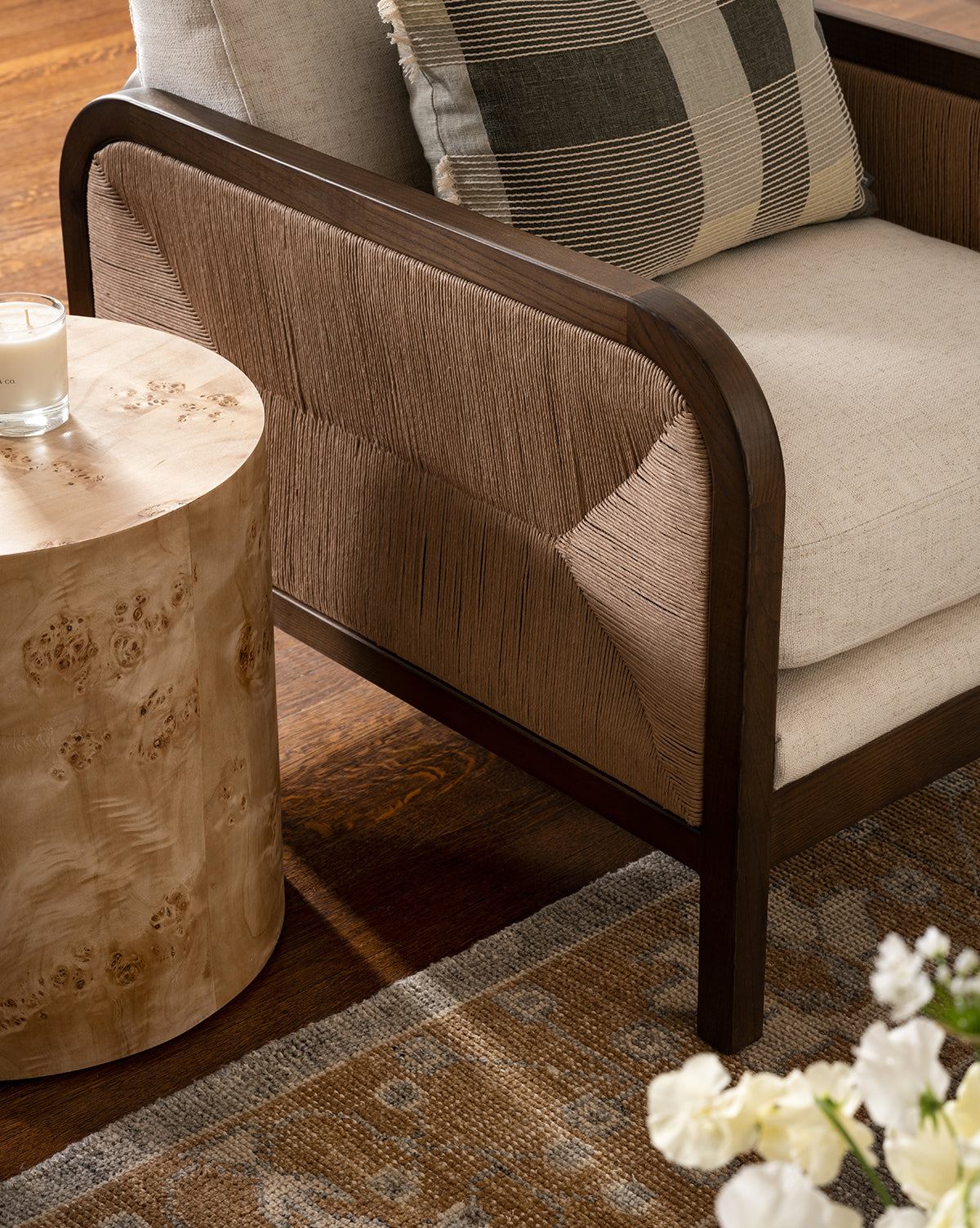 A close-up of the Garvey Lounge Chair with woven sides, beige upholstered cushions, and a plaid pillow beside a solid ash round side table with a candle. Partially visible are a patterned rug and white flowers, by McGee & Co.