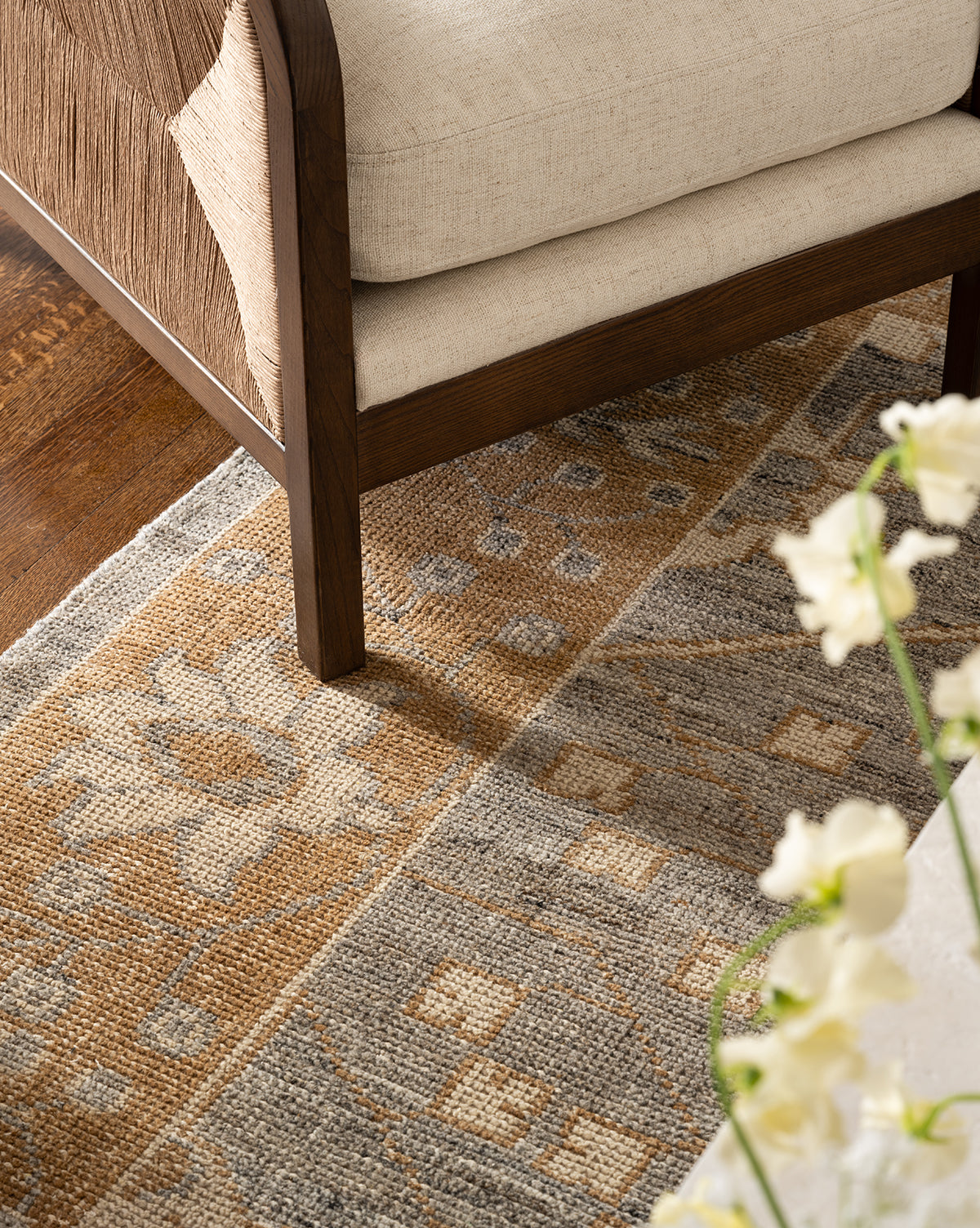 A close-up of a beige cushioned wooden chair on the Elison Hand-Knotted Wool Rug featuring gray, tan, and cream tones; white flowers are partially visible in the foreground, by McGee & Co.
