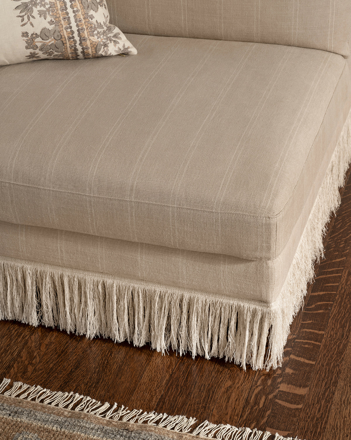 The Philippa Settee (Ready to Ship) in beige features decorative fringe along the bottom edge, set on a dark wooden floor. A patterned pillow rests on the settee and part of a woven rug is also visible, by McGee & Co.