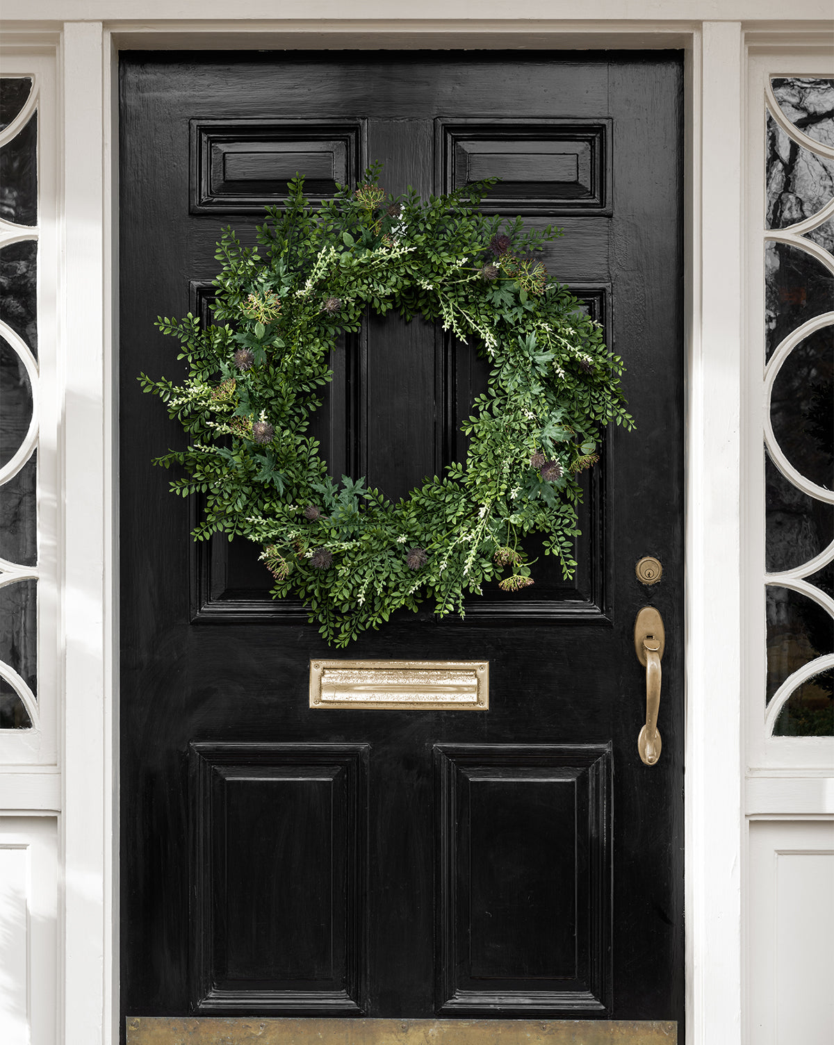 A large Faux Summerset Wreath made of lush artificial greenery hangs on a black front door with white trim, brass mail slot, and door handle. Circular window designs complement this elegant seasonal decor.