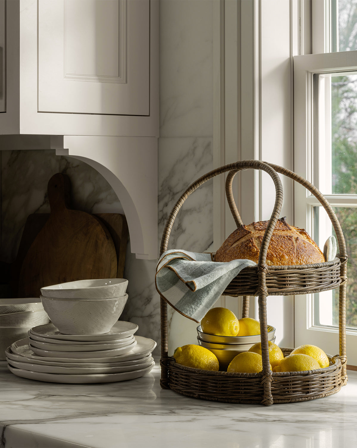 The Aneesa Two-Tiered Wicker Tray, filled with a loaf of bread, a folded cloth, and lemons, rests on a marble kitchen counter near stacked white dishes by a sunlit window by McGee & Co.