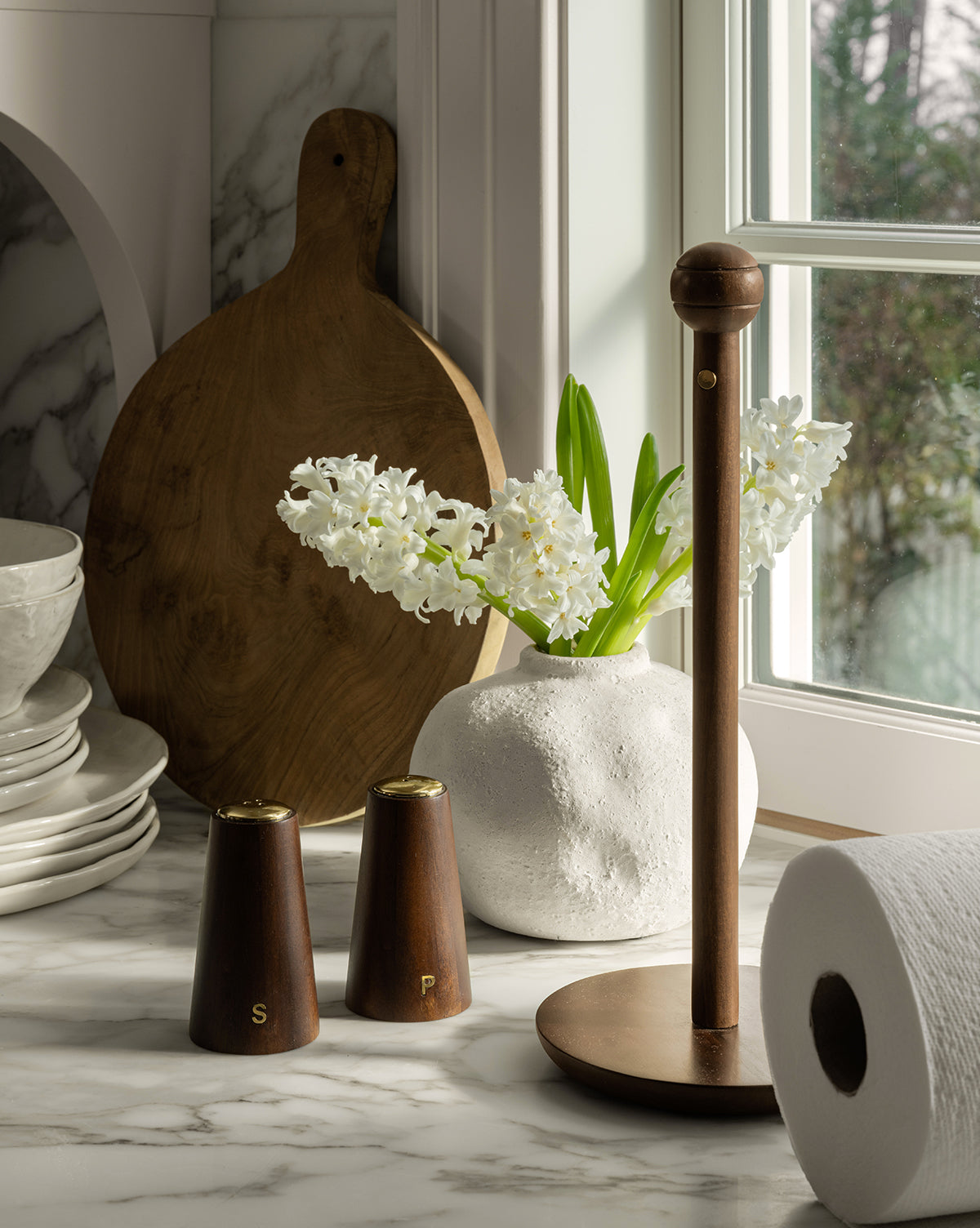 A marble kitchen countertop with French café charm features a white vase of hyacinths, Brasserie Salt & Pepper Shakers, cutting boards, stacked dishes, a wooden paper towel holder, and a roll of paper towels by a bright window.