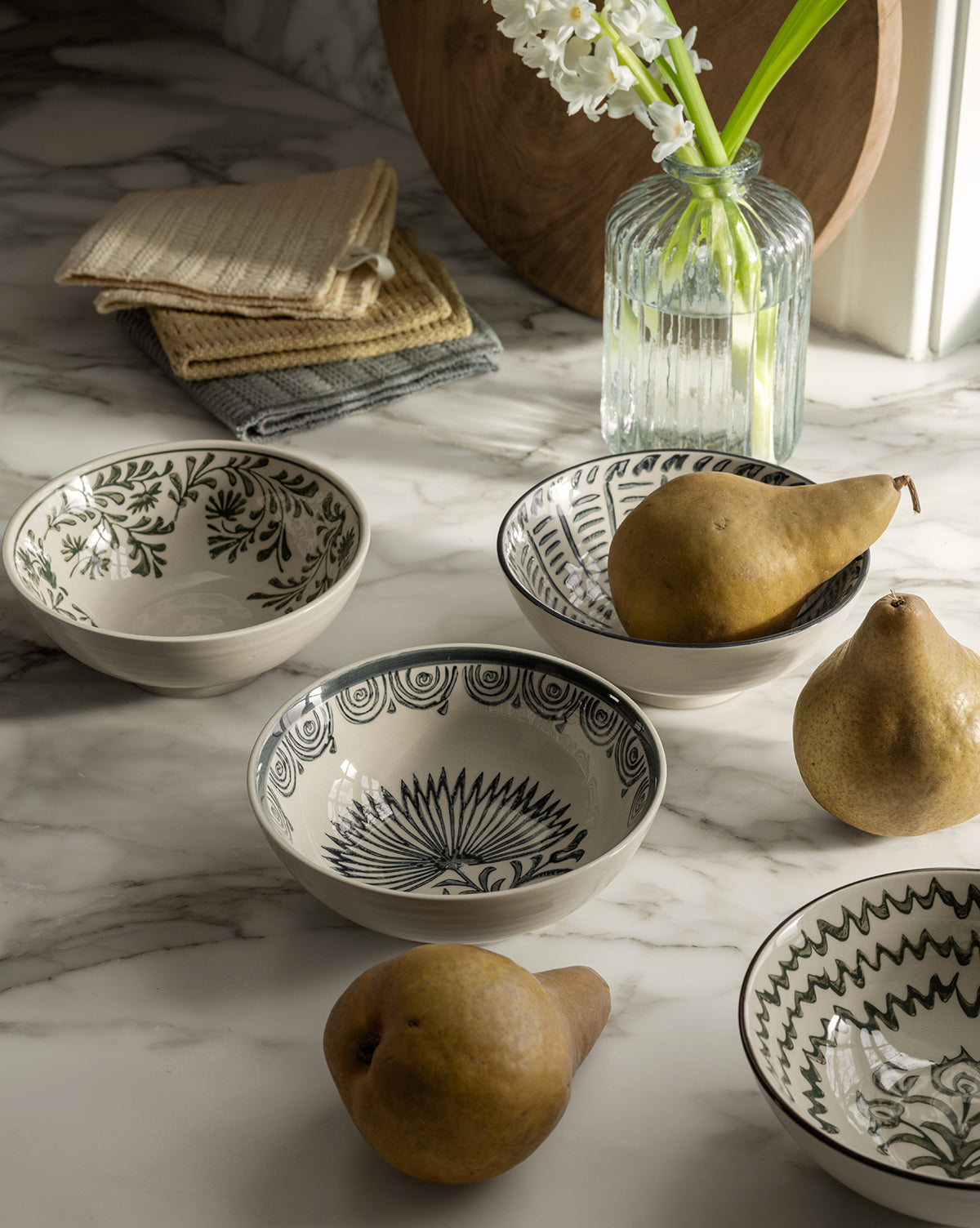 Four glazed stoneware bowls with floral patterns and three pears rest on a marble countertop. A Mattia Hand-Painted Bowl, white flowers in a glass vase, and stacked folded cloths are arranged near a window in the background by McGee & Co.