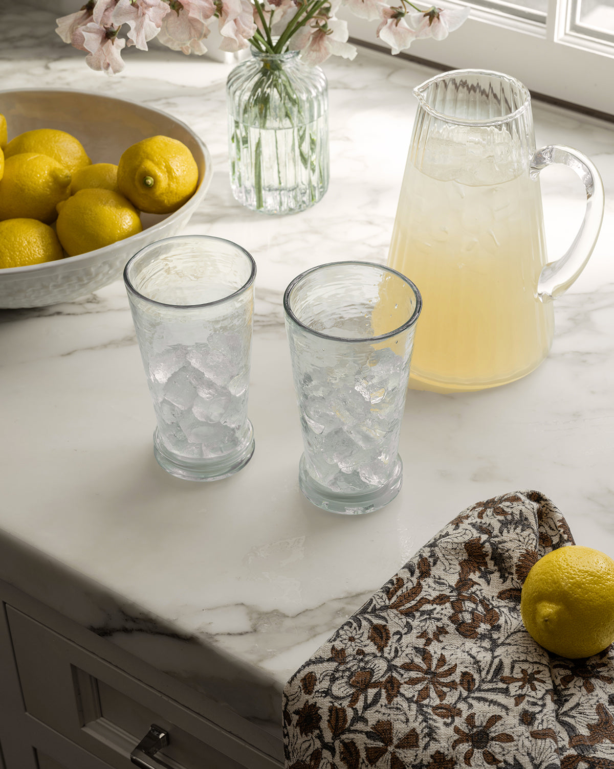 The Adonis Ribbed Glass Pitcher, filled with lemonade and paired with ice-filled glasses, fresh lemons, flowers, and a patterned cloth on a sunlit marble countertop, creates an effortlessly stylish kitchen scene, by McGee & Co.
