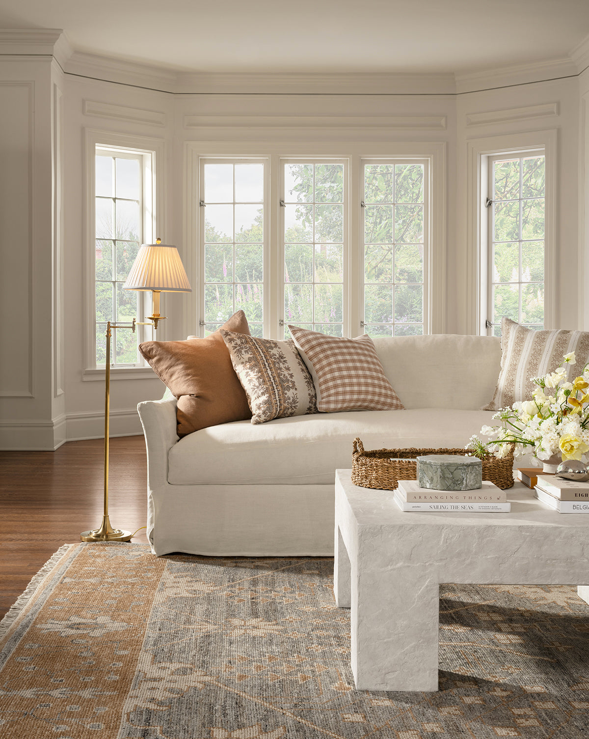A bright living room featuring the Wilhelmina Classic Slipcover Sofa (85-110) with brown and plaid pillows, a marble coffee table with books and flowers, a brass floor lamp, and large bay windows overlooking greenery by McGee & Co.