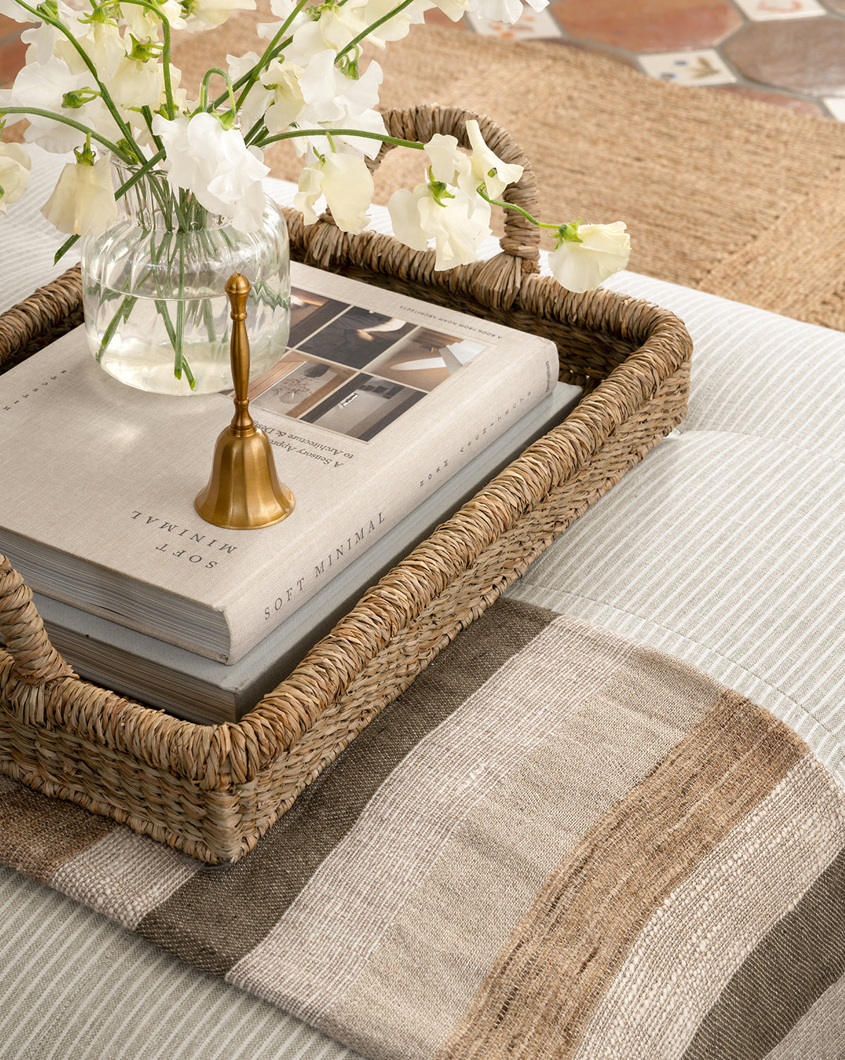 A woven tray holds a glass vase with white flowers, a book titled Soft Minimal, and a gold candle snuffer on light striped fabric, with the Ashwood Throw—a neutral-toned decorative accent—nearby.