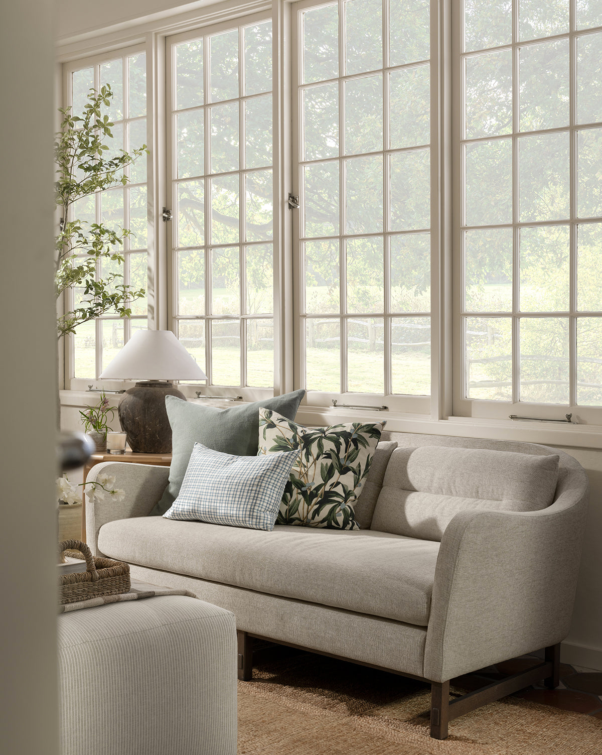 The Commodore Settee 68 in light gray twine crypton upholstery, with patterned and solid pillows, sits by large windows in a bright living room with greenery, a table lamp, and neutral decor as sunlight streams through the trees outside, by McGee & Co.