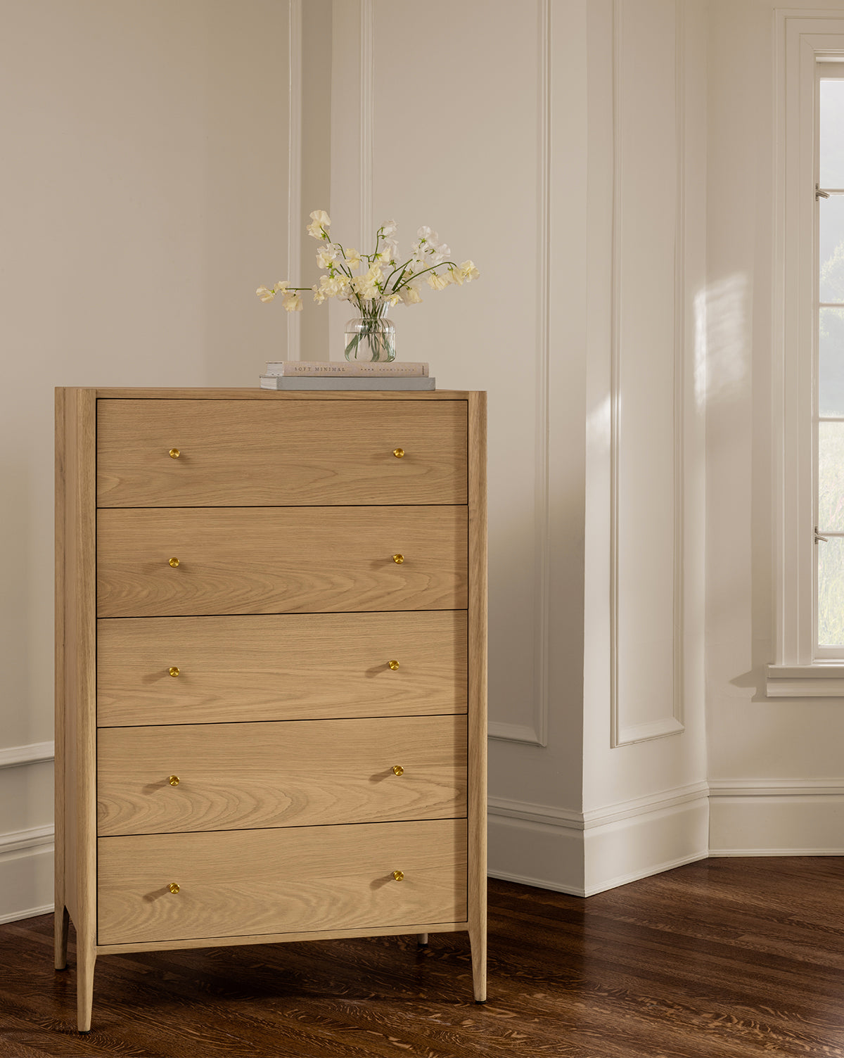 The Dana Tallboy Dresser in white oak with five drawers and brass hardware stands on a hardwood floor. Stacked books and a glass vase with white flowers sit on top, bathed in sunlight from a window on the right, by McGee & Co.