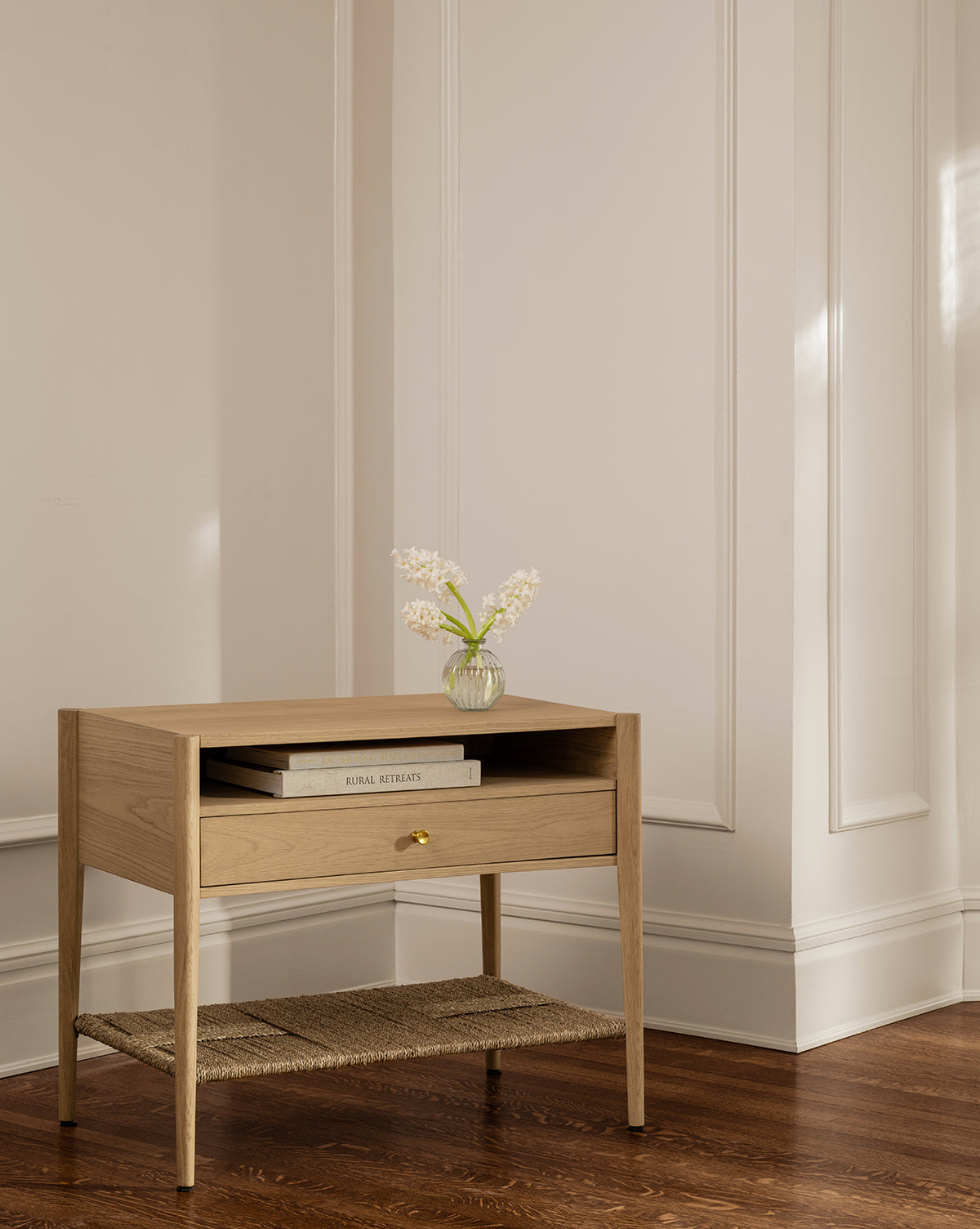 The Dana White Oak Nightstand, featuring a single drawer and woven seagrass shelf, is placed against a cream wall with two stacked books and a small glass vase of white flowers on top, gently lit by sunlight by McGee & Co.