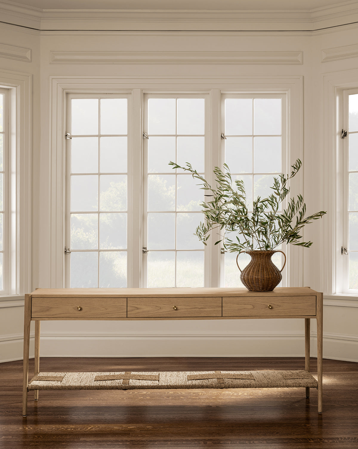 The Dana White Oak Console with three soft-close drawers stands before white-trimmed windows. A brown ceramic vase with leafy branches sits on top. The space features hardwood floors and neutral-colored walls by McGee & Co.