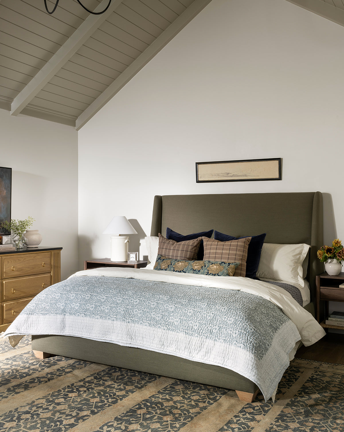 A cozy bedroom features a green upholstered bed with blue and plaid pillows and an artisanal bedding set, topped by the Blue Floral Block Print Quilt. Wooden nightstands hold lamps and flowers, with a dresser left under the vaulted gray ceiling, by McGee & Co.