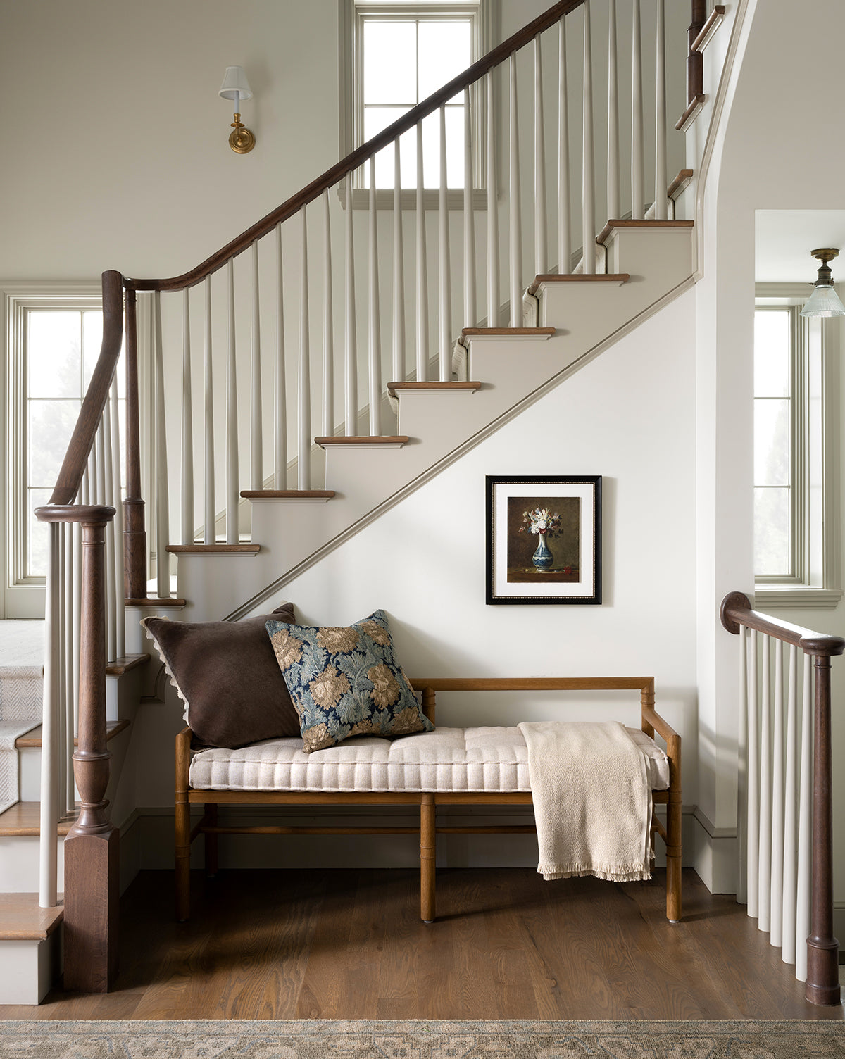 A wooden bench with cushions and a throw blanket sits beneath a staircase, while vintage-inspired wall art featuring the Antique Flower Vase hangs above, illuminated by natural light from nearby windows, by McGee & Co.