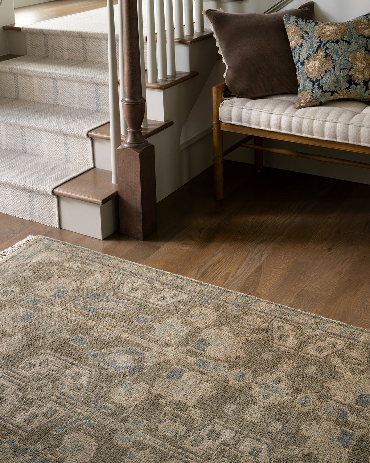 The Chevalier Hand-Knotted Wool Rug in neutral tones lies on the wooden floor near a staircase with carpeted steps. A wooden bench with a beige cushion and pillows beside the stairs adds timeless elegance to the space, by McGee & Co.