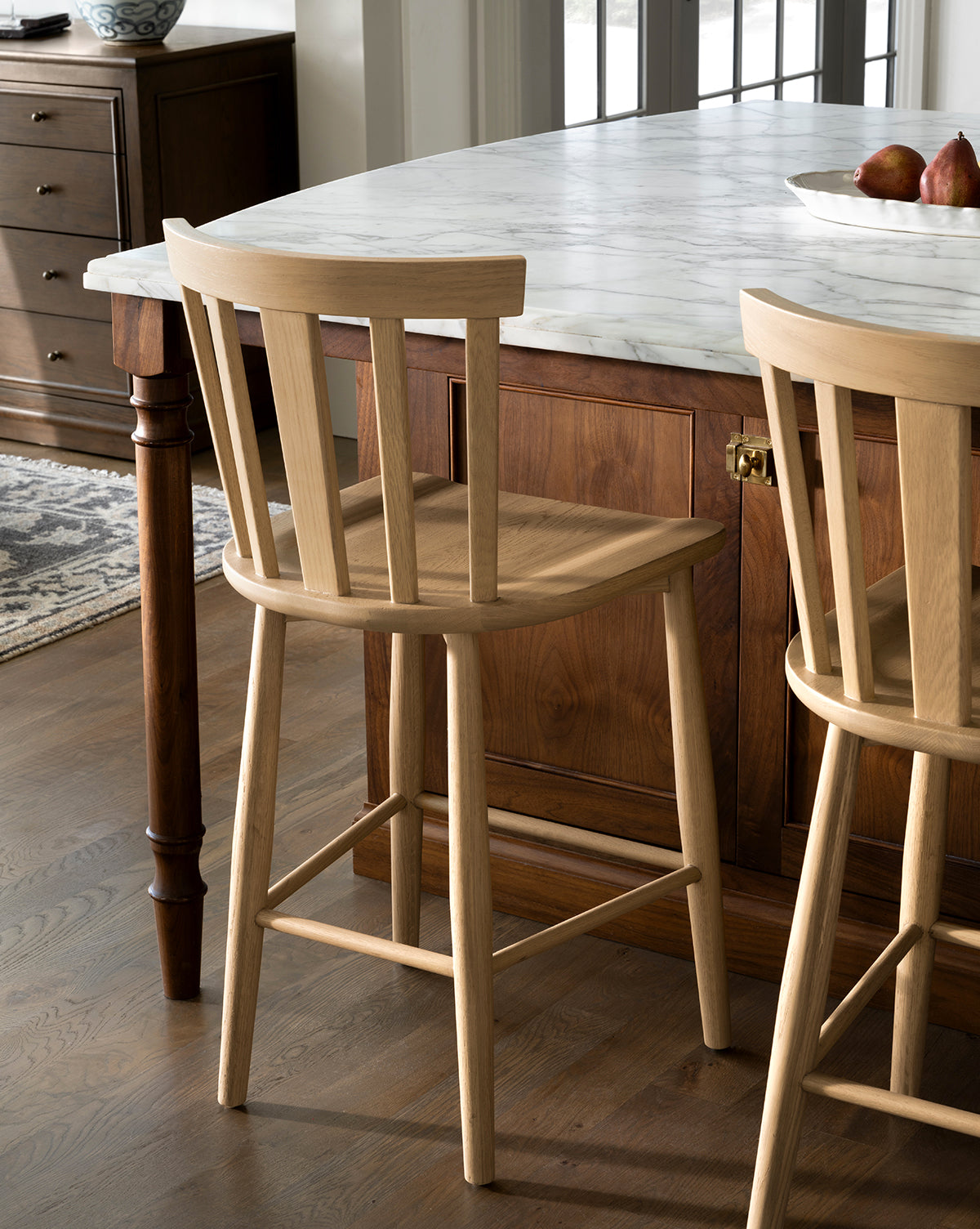 A Hemingway Stool made from solid wood with a slatted backrest stands by a marble-topped kitchen island, complementing the wooden floor and cabinetry. A second Hemingway Stool is partially visible near the kitchen counters, by McGee & Co.