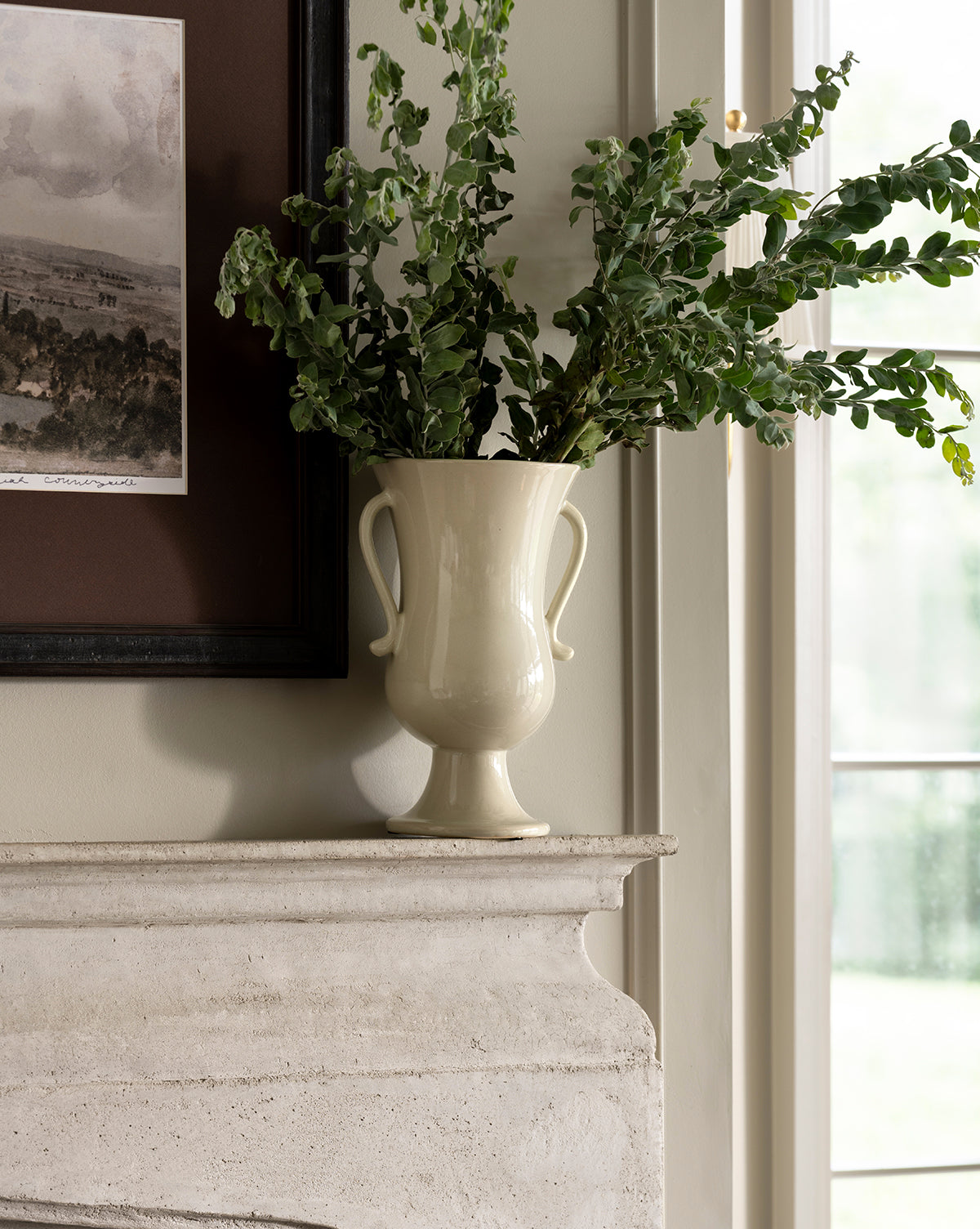 The Delphine Footed Vase, cream-colored and elegant with two handles, holds leafy green branches on a stone mantel beside a framed picture and a sunlit window, by McGee & Co.