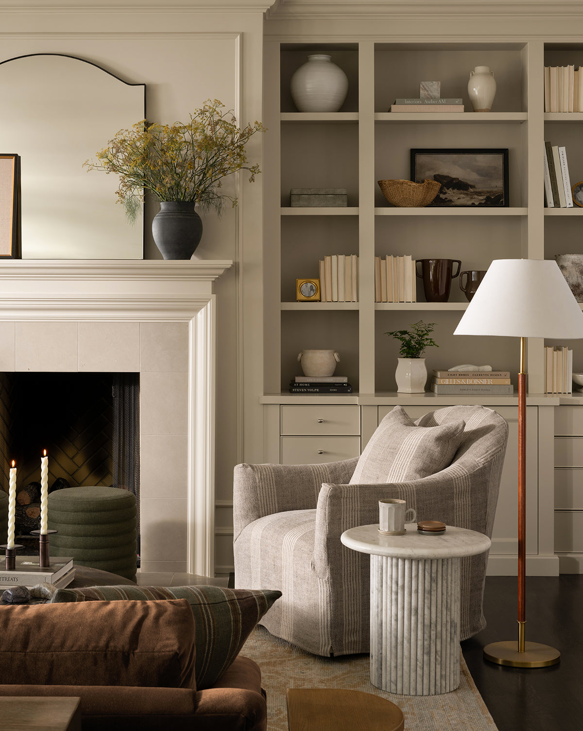A cozy living room features a light armchair by a round marble side table and the Elijah Floor Lamp with a brass pole. Built-in shelves display books, vases, and baskets. To the left is a fireplace with candles beside a brown sofa with leather accents, by McGee & Co.