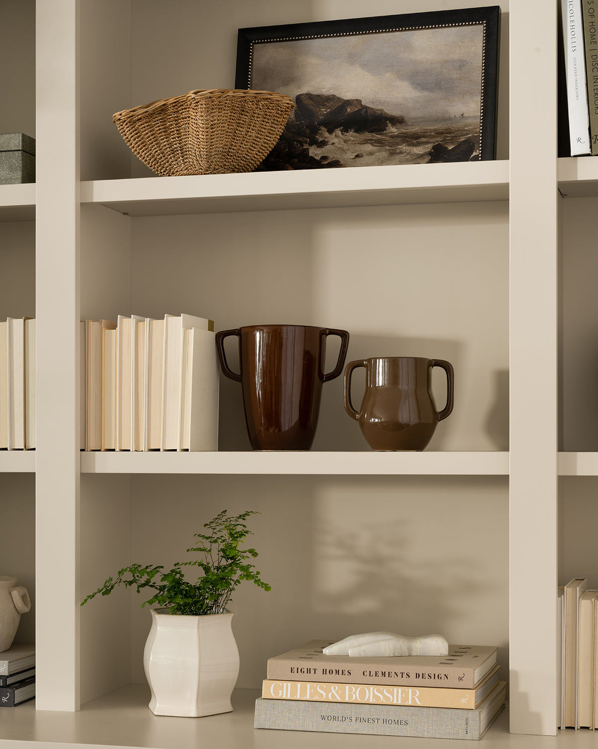 A beige bookshelf displays a basket, a gallery-quality framed print of Rough Sea Shores, two brown handled vases, a white vase with greenery, and stacks of books arranged both vertically and horizontally by McGee & Co.