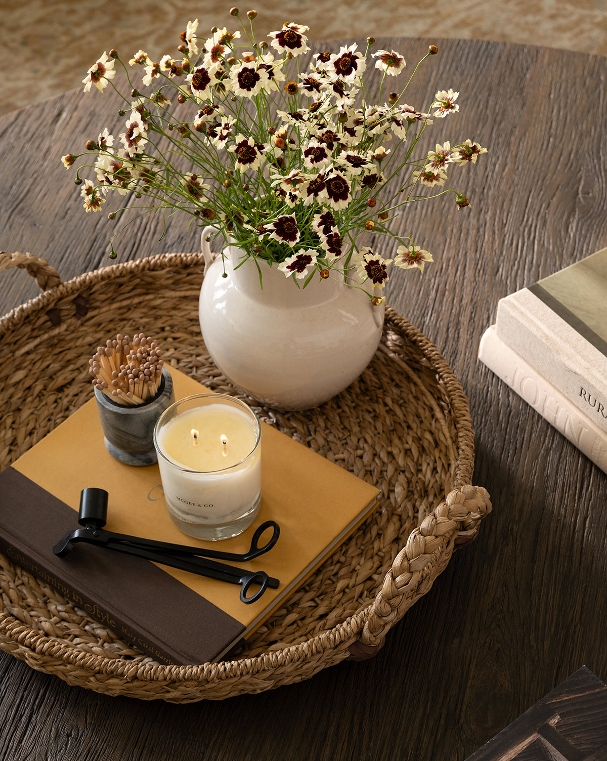 A woven tray on a wooden table holds a white vase with small white and dark red flowers, a lit candle, a match holder, the Wick Trimmer for candle care, and two books; nearby is another stack of books.