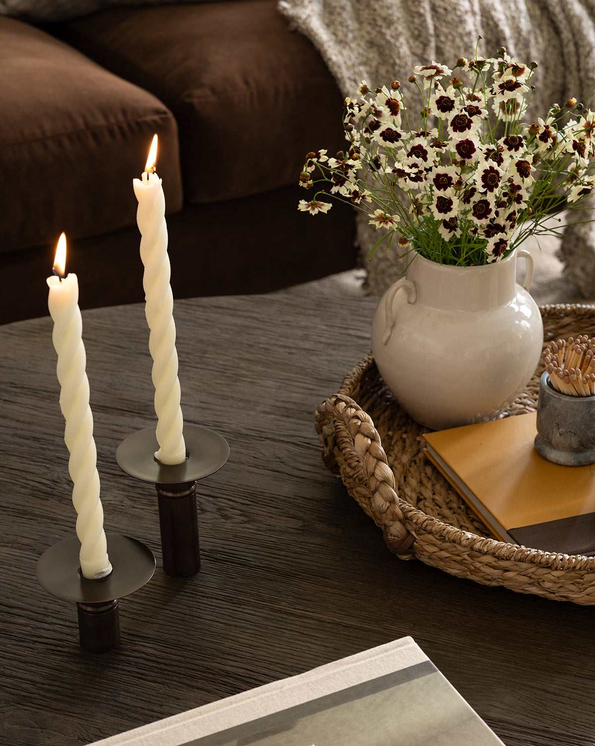Two twisted white candles in black holders are lit on a dark wooden table. Nearby, a woven tray holds the Rasley Handled Jar with white glaze, a vase of daisies, and stacked notebooks. A brown couch with a textured throw blanket is in the background, by McGee & Co.
