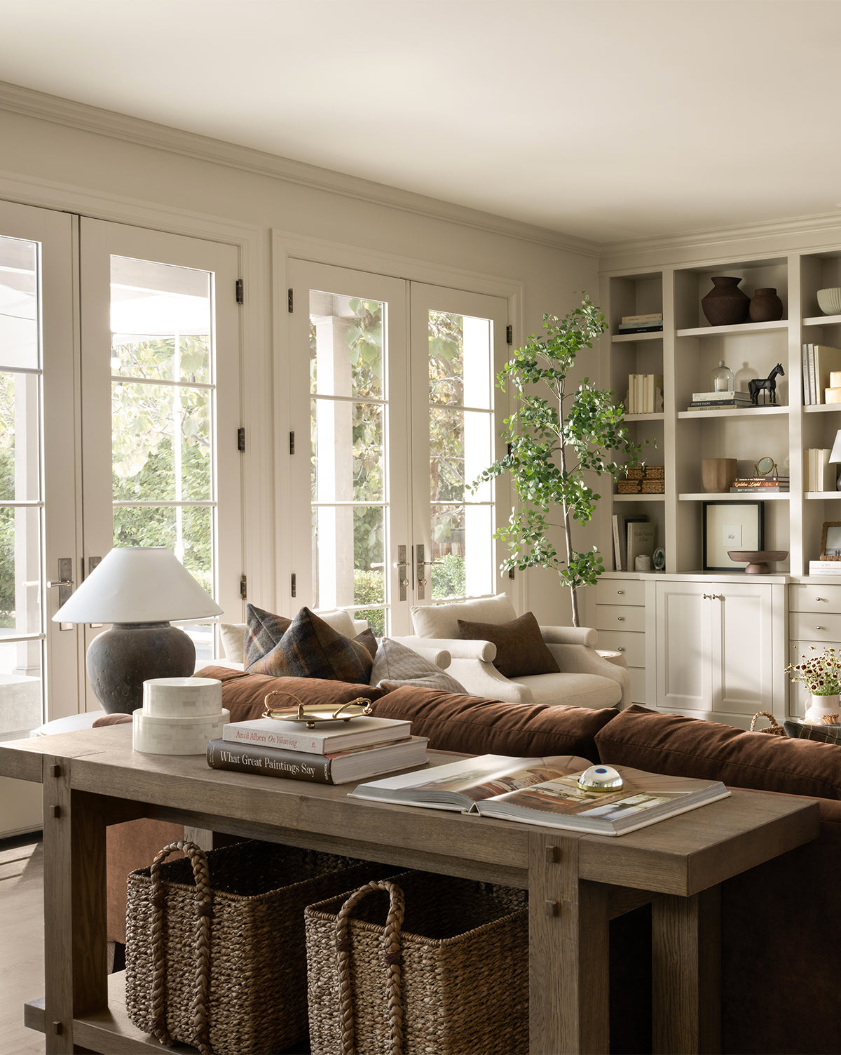 A cozy, sunlit living room with French doors, a brown sofa, and a wooden console table featuring the Salter Woven Square Basket, books, and decor; built-in shelves display vases, books, and plants for a warm, inviting space by McGee & Co.