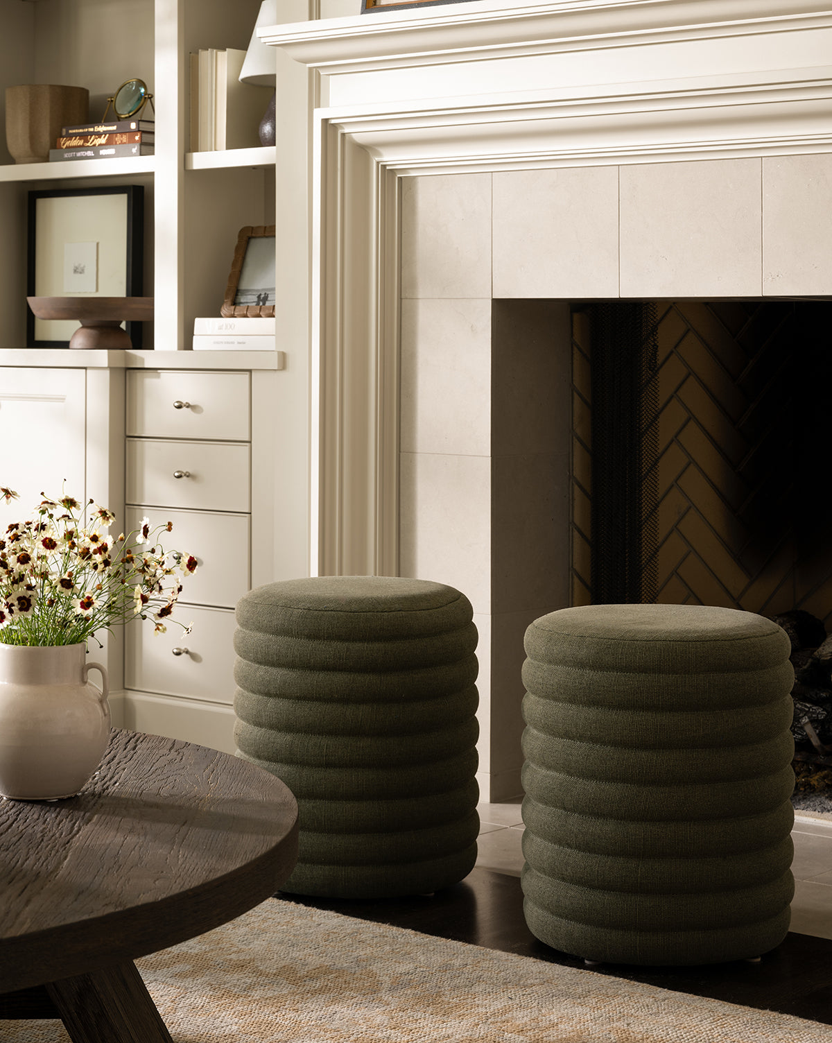 Two round, textured Jefferson Stools by Made Goods in green quilted fabric sit before a white-mantel fireplace. A wooden coffee table with a flower vase and built-in shelves/cabinets can be seen in the background, blending seamlessly with touches from McGee & Co.