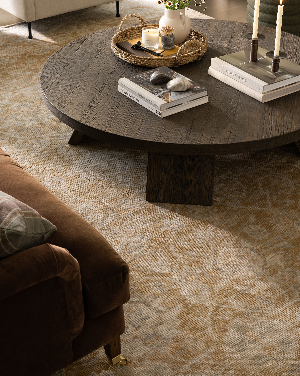 A round, dark wood coffee table topped with books, stones, candles, and a wicker tray sits on the Vanderwood Hand-Knotted Wool Rug near a brown upholstered sofa and cream armchair in a cozy living room, by McGee & Co.