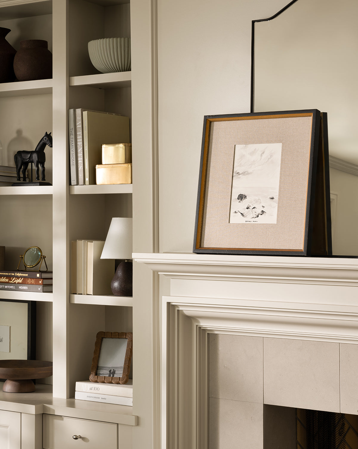 A neutral-toned living room features built-in shelves and decor, with September in Kalamata—a giclee print with a hand-wrapped linen mat in a distressed black frame—displayed above the fireplace and reflected in the mirror behind it.
