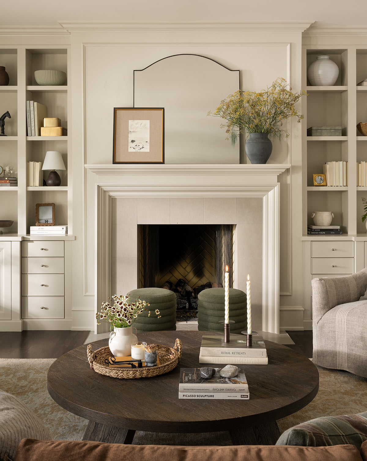 A cozy living room features a fireplace, built-in shelves, and a round dark wood coffee table with books, candles, and a vase. The Willson Wall Mirror with its iron frame enhances the decor as neutral tones and soft lighting create warmth by McGee & Co.