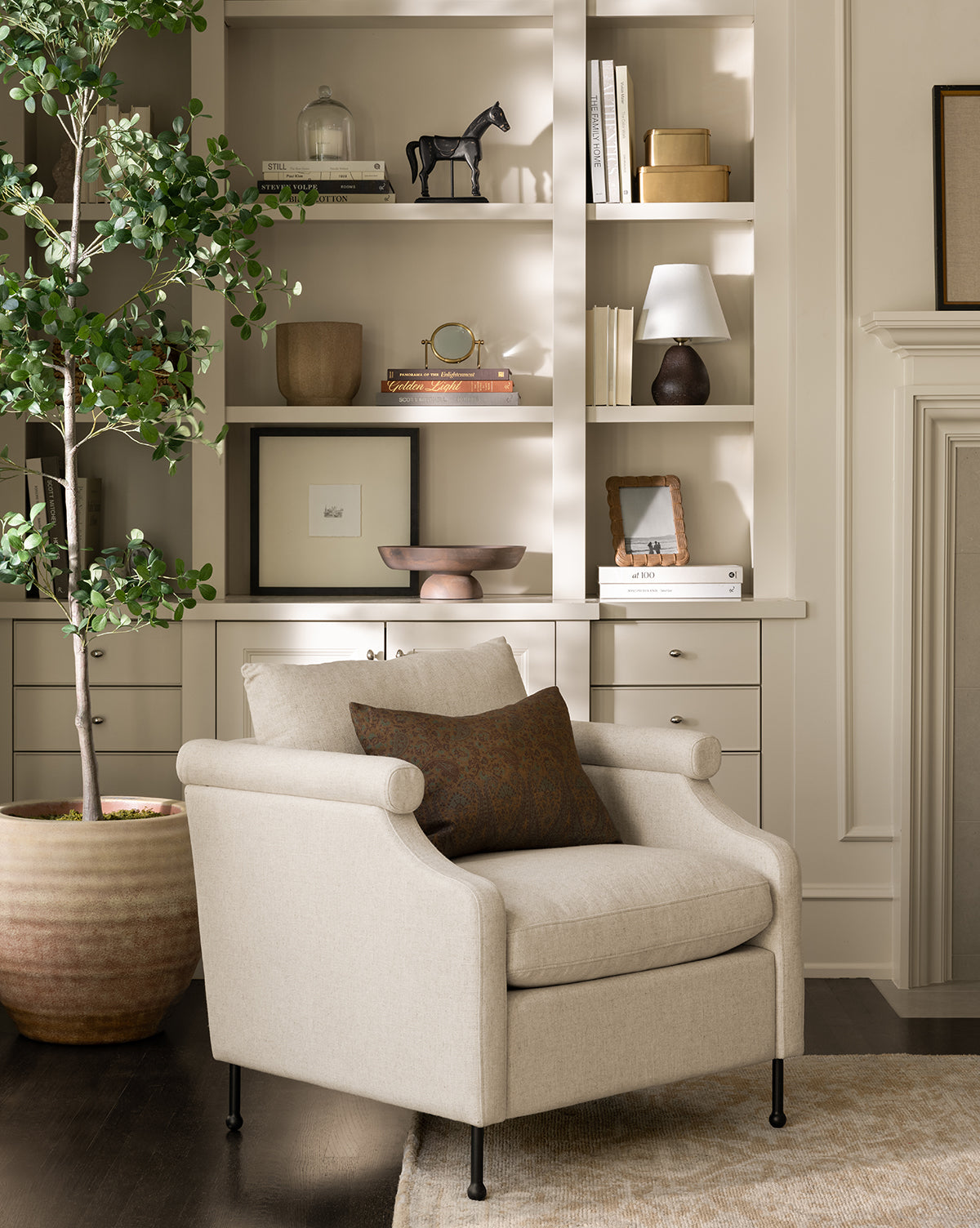 A cozy beige Clegg Lounge Chair by Makerspalm with a brown pillow sits before built-in shelves of books, decor, and a lamp. A large potted plant on the dark wood floor brings vintage charm to this inviting living space. Styled with accents from McGee & Co.