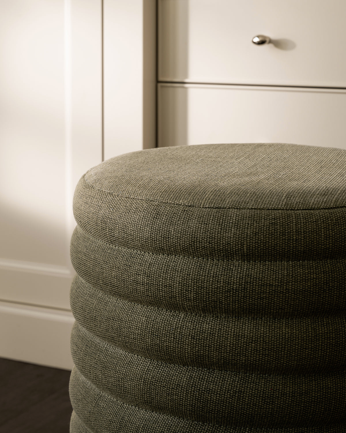 A close-up of the Made Goods Jefferson Stool, featuring a round, textured gray quilted design with horizontal ridges, positioned in front of light-colored cabinets and drawers with metallic knobs. McGee & Co.
