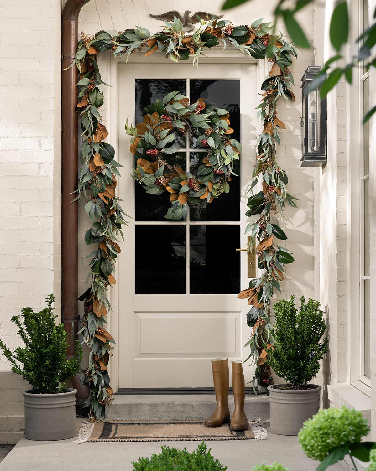 A cream-colored door is adorned with a Faux Magnolia & Eucalyptus Garland and wreath, while two potted plants and tan boots sit on the doormat, creating a warm welcome framed by light brick walls, by McGee & Co.