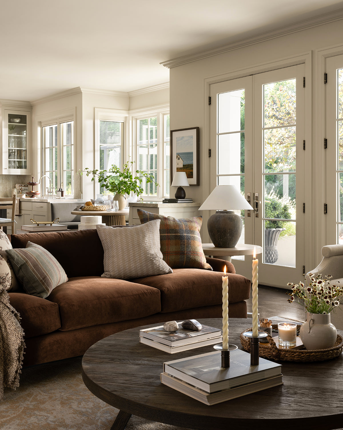 A cozy, sunlit living room features a customizable Lucille English Roll Arm Sofa 90.5 with down wrapped cushions and patterned pillows, a round wooden coffee table with candles and books, large windows, French doors to the garden, and the kitchen in view, by McGee & Co.