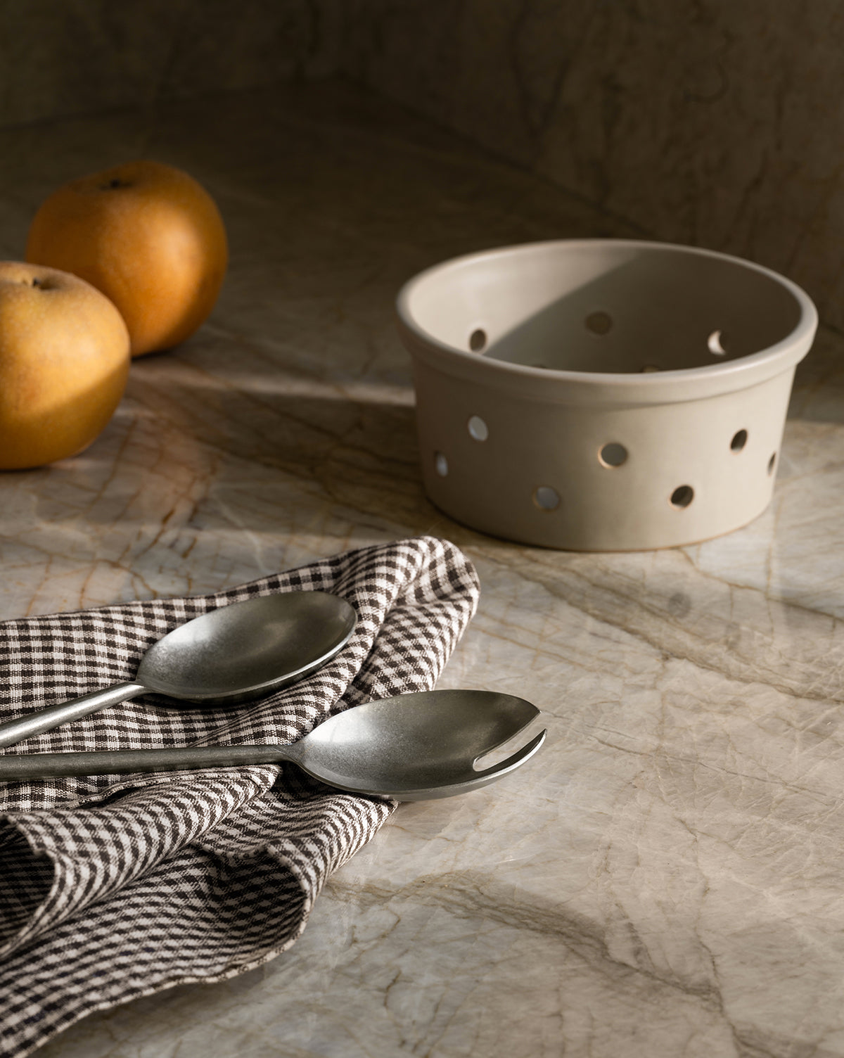 Two metal spoons rest on the Westport Linen Tea Towel atop a marble surface. Warm natural light casts soft shadows, highlighting the towel alongside two apples and a beige ceramic basket with holes, by McGee & Co.