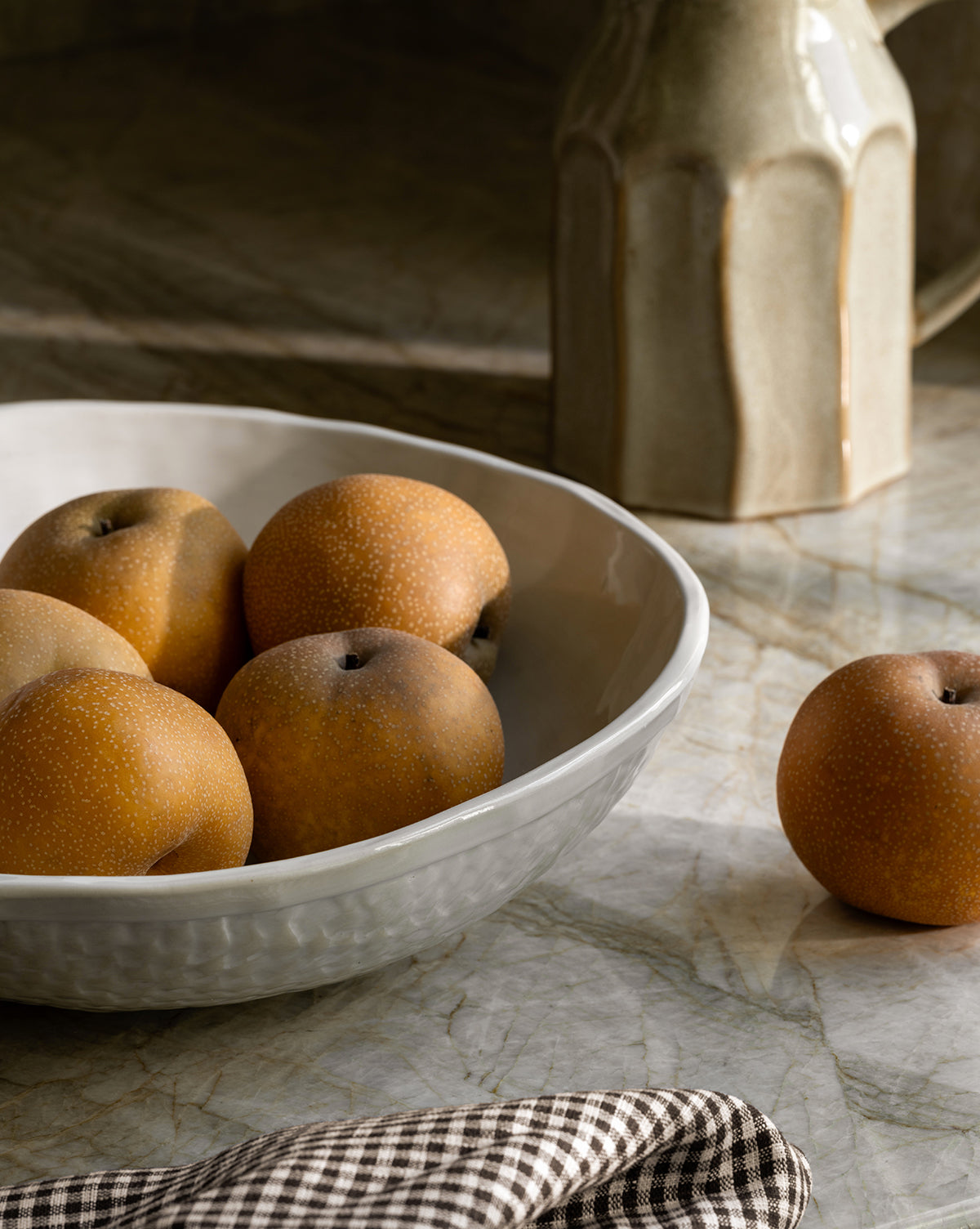 The Dion Serving Bowl, finished in white ceramic glaze, holds five Asian pears on a marble surface with another pear nearby. A beige jug and checked cloth add elegance to the tranquil arrangement by McGee & Co.