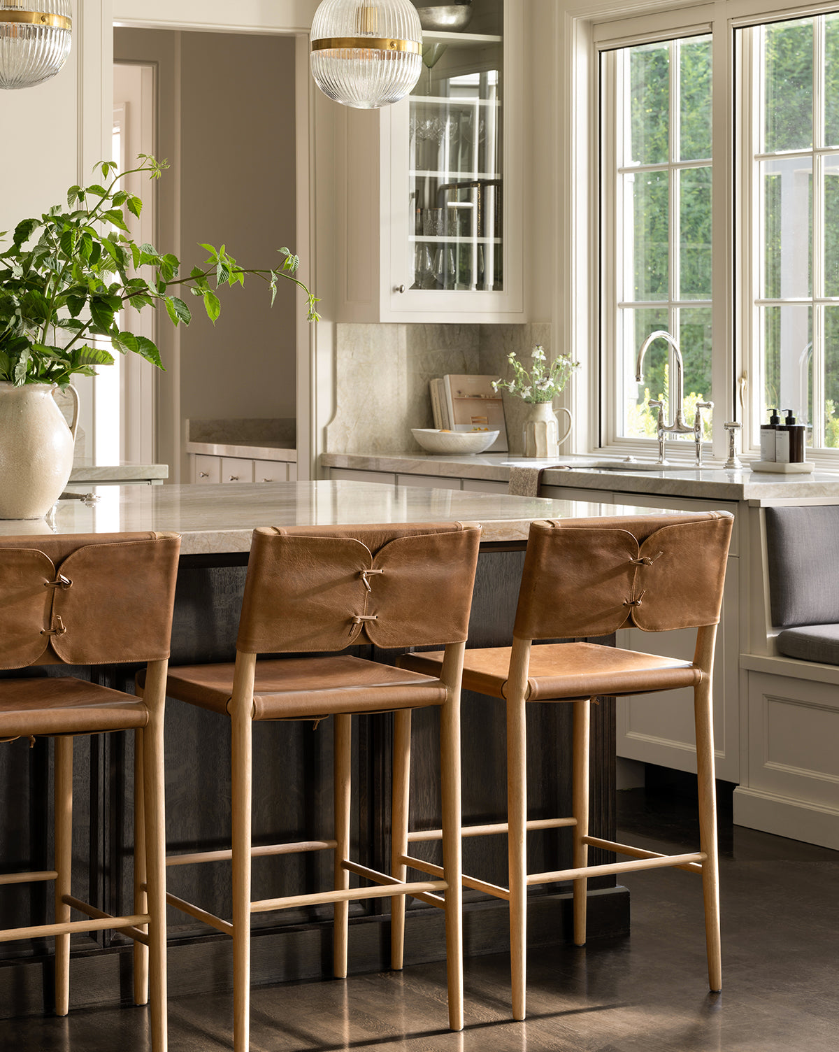 A modern kitchen showcases Danson Counter Stools with tan leather seats at a marble island, large windows bringing in natural light, minimalist neutral cabinetry, and a potted plant by McGee & Co.