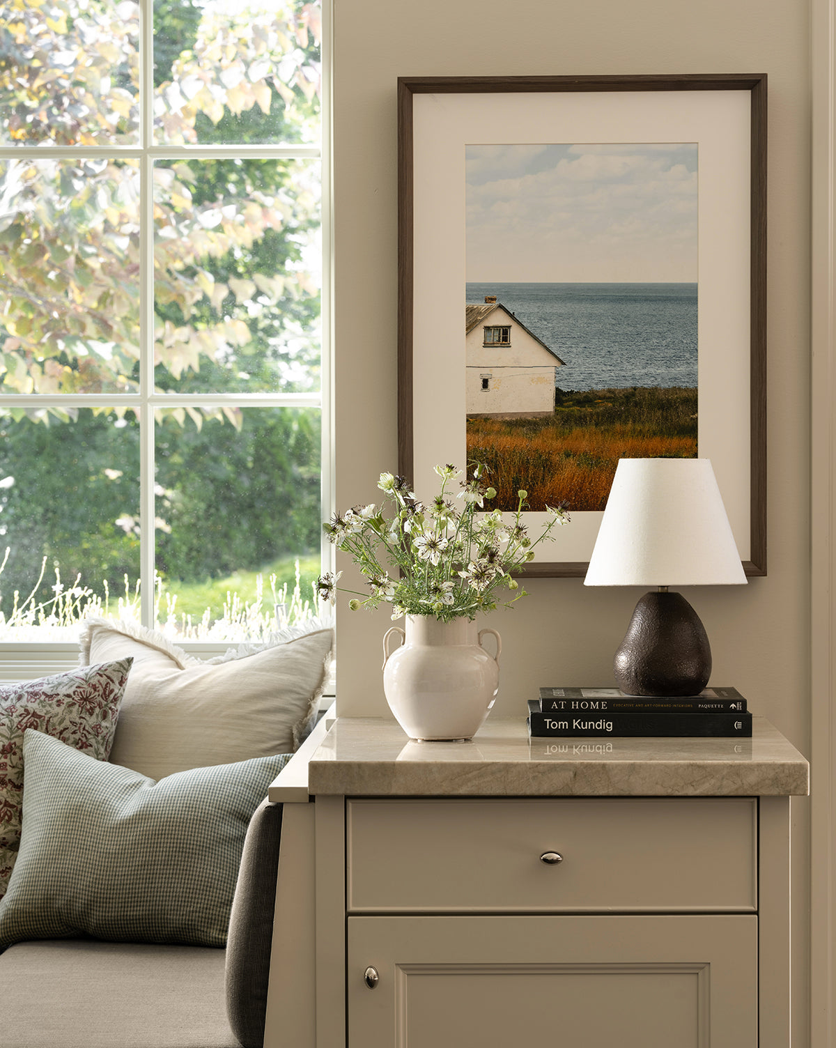 A cozy reading nook with neutral cushions sits by a sunlit window. On a small cabinet are decor items, while Room with a View, a gallery-quality art piece in a dark walnut frame, hangs on the wall above, by McGee & Co.