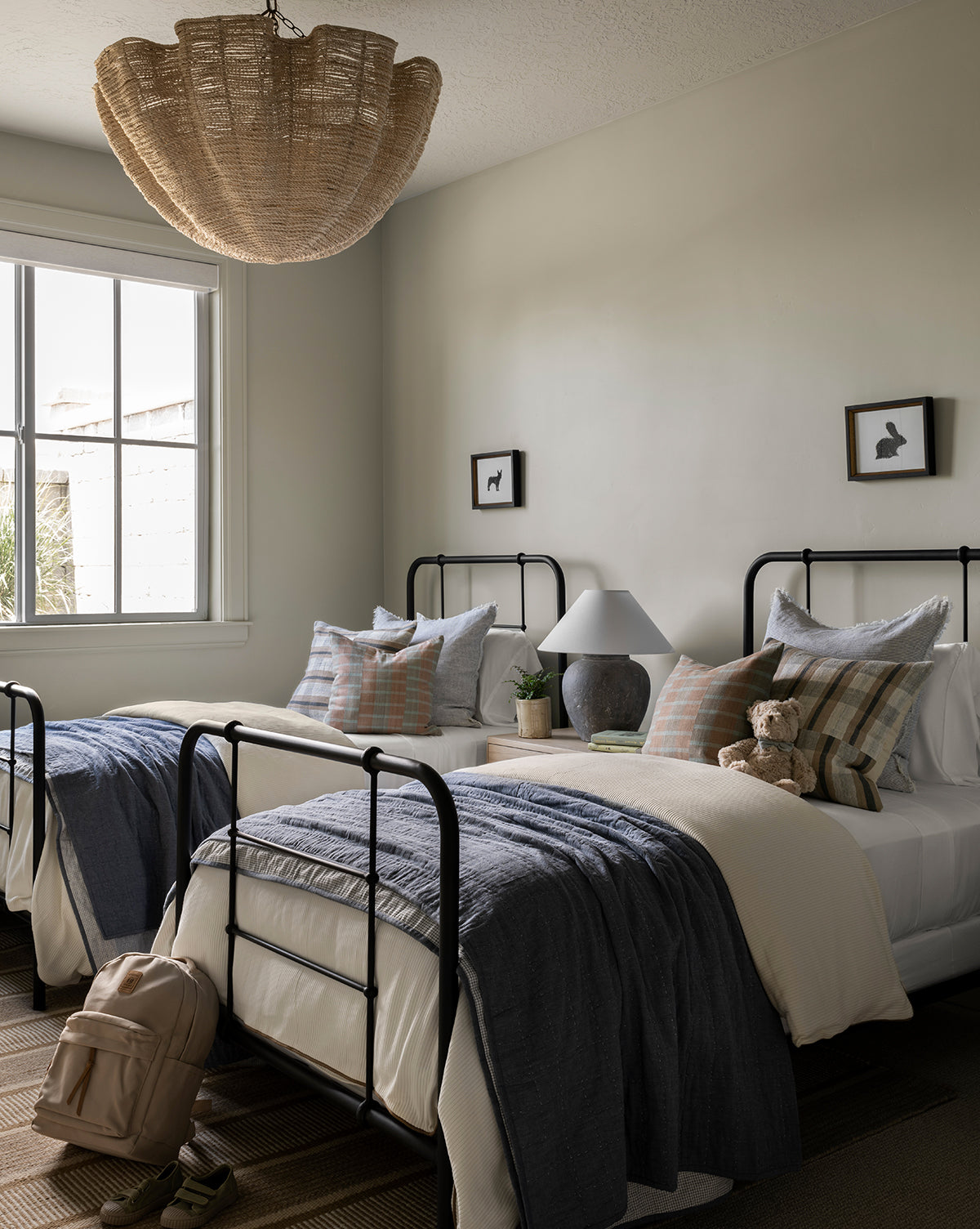 Two twin beds with black metal frames are side by side in a cozy, neutral-toned room. Each bed is topped with the Fitzgerald Kids Quilt and decorative pillows, while a woven light fixture hangs above and a backpack sits at the foot of one bed.