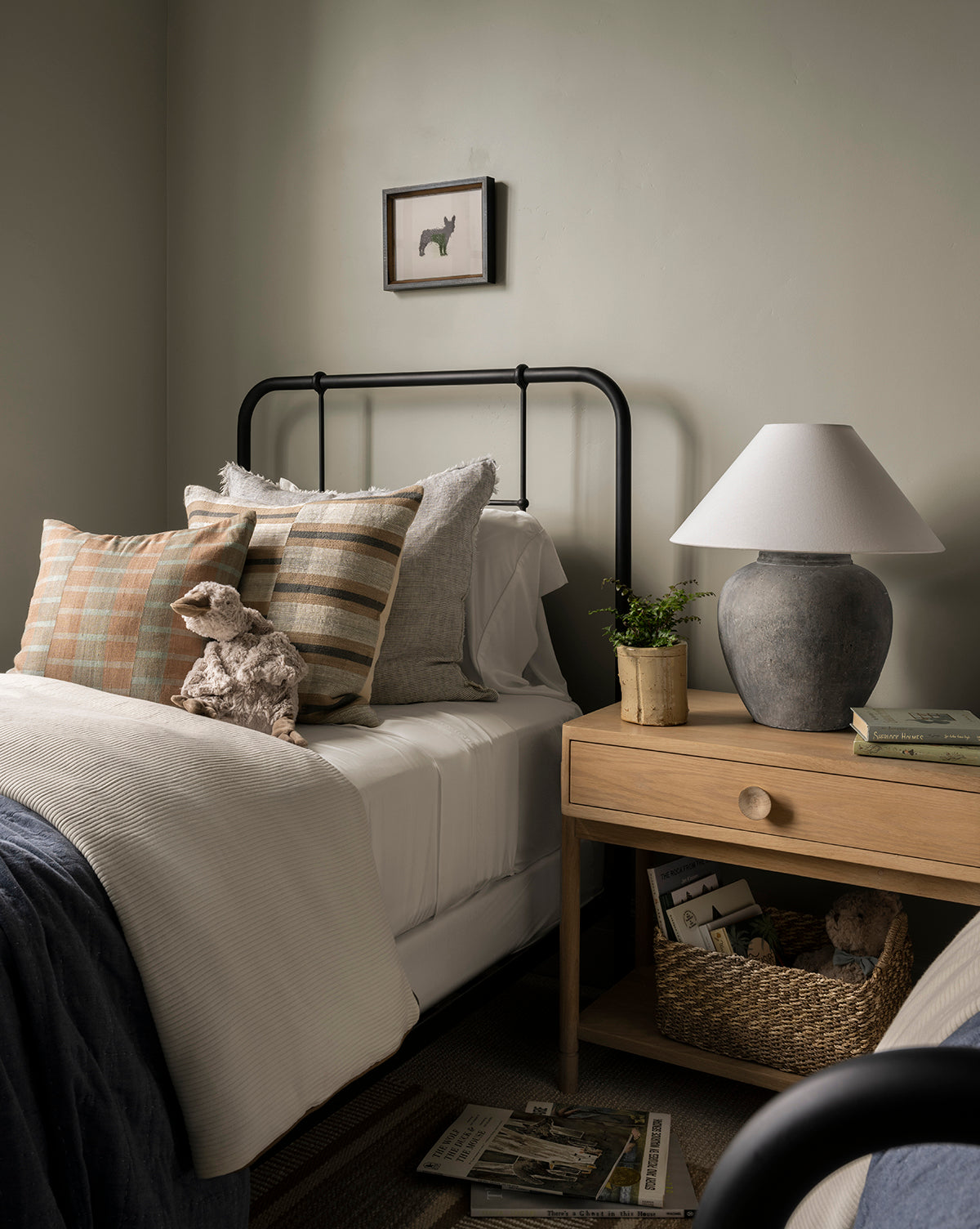 A cozy bedroom with a black metal bed, neutral pillows, a plush dog toy, and a wooden nightstand featuring the Montague Table Lamp, books, and a potted plant. A framed animal print hangs above the bed, by McGee & Co.