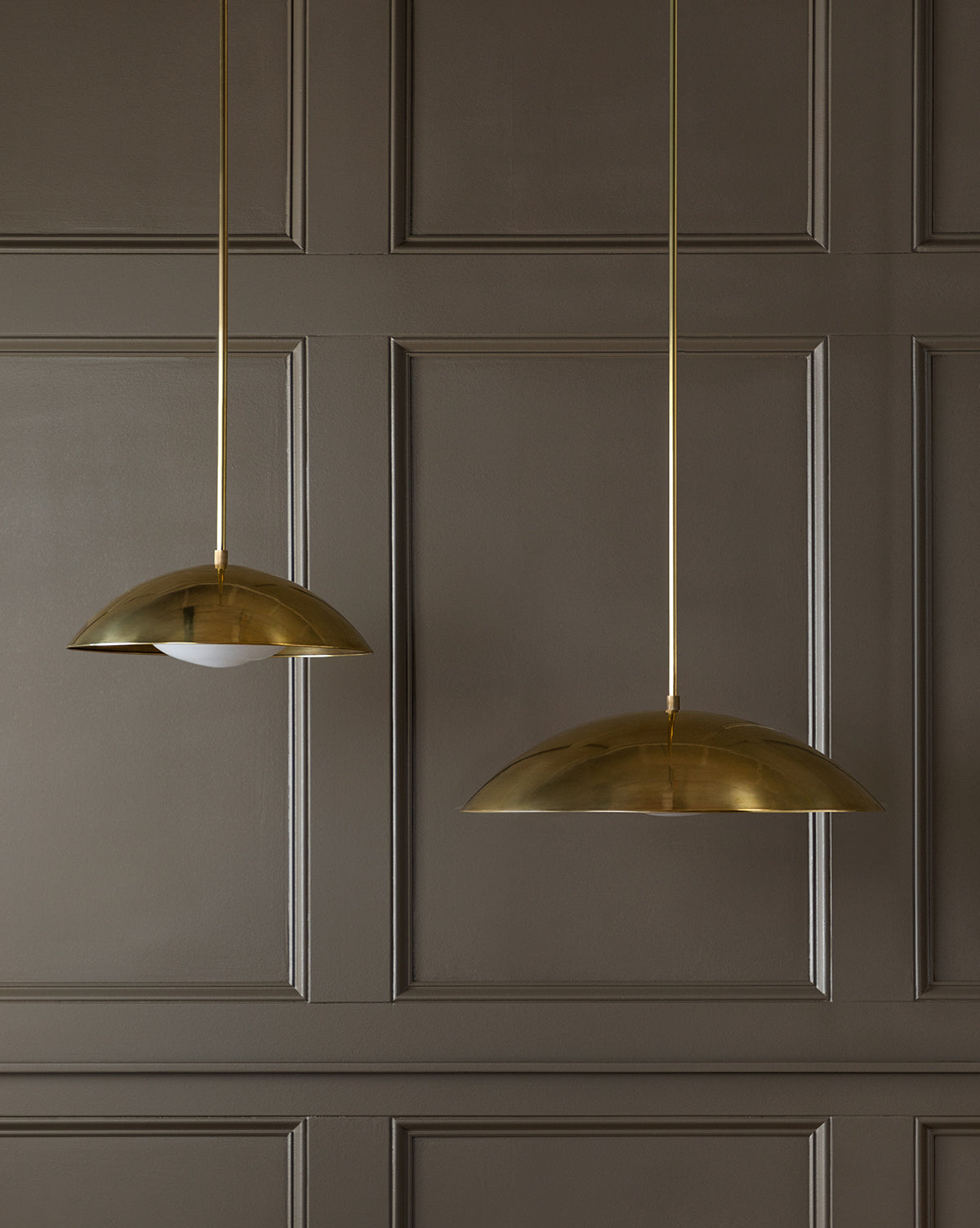 Two modern brass Aldorno Pendants hang before a brown paneled wall: the left exposes its bulb, while the right showcases a wide, round shade that fully conceals the bulb, by McGee & Co.
